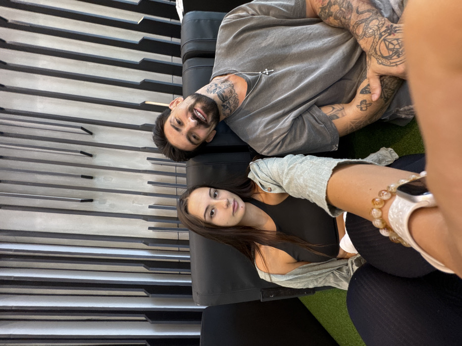 An indoor selfie-style photograph captures two individuals, a female and a male, seated side-by-side in black lounge chairs. The female, positioned on the left and holding the camera, has long dark hair, a neutral expression, and wears a black crop top, an unbuttoned light green long-sleeved shirt, and dark trousers. A white Apple Watch and a beaded bracelet are visible on her right wrist. The male, on the right, has short dark hair, a beard, extensive tattoos on his neck and arms, and is smiling directly at the camera with his head tilted slightly. He wears a grey t-shirt and a silver chain with a cross pendant. The background features a modern interior wall with a repetitive pattern of vertical, dark-colored, abstract slat designs against a lighter grey surface. The floor beneath them is covered in green, artificial turf-like material. The precise location within Bangkok, Thailand, is not discernible from the visual cues, but the environment is consistent with a contemporary commercial establishment or waiting area.