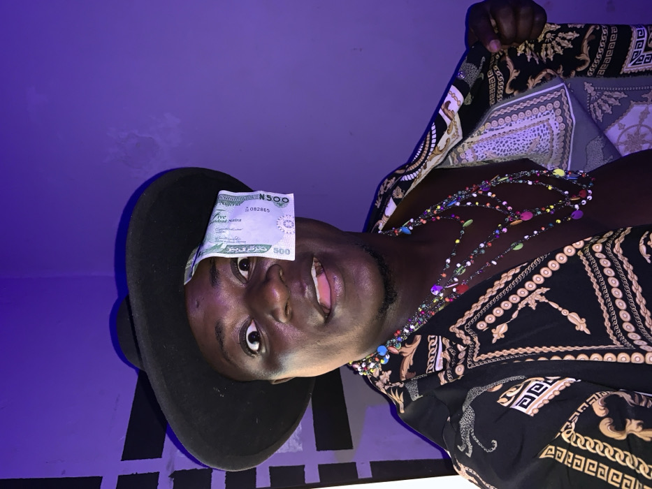 A man smiles at the camera wearing a black hat with a 500 Nigerian Naira bill stuck to it. He wears an open, patterned shirt with a silver and gold design, revealing his dark skin and colorful beaded necklaces. The lighting is dim and tinted blue. A black and white striped wall is behind him to the left, and his hand is at the top right.