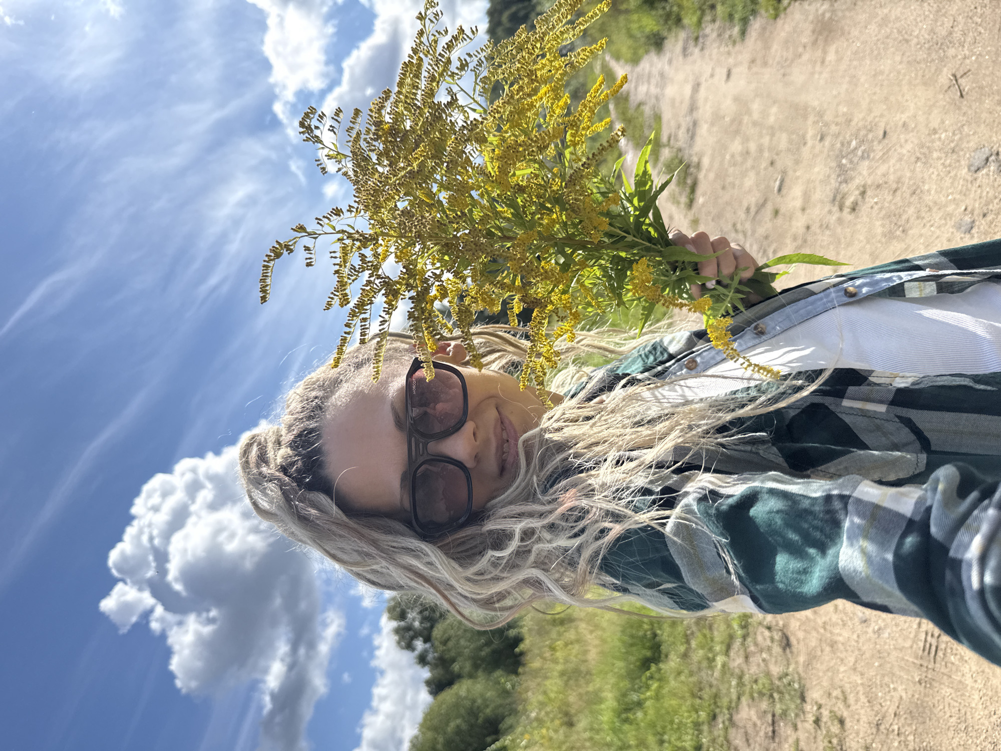 A blonde woman with her hair in a high ponytail and wearing sunglasses and a green and black plaid shirt holds a bouquet of yellow wildflowers. She is standing on a dirt path in a grassy field, with trees in the background. The sky is blue with white clouds. The image was taken outdoors. This scene is in Rusanovo, Russia.