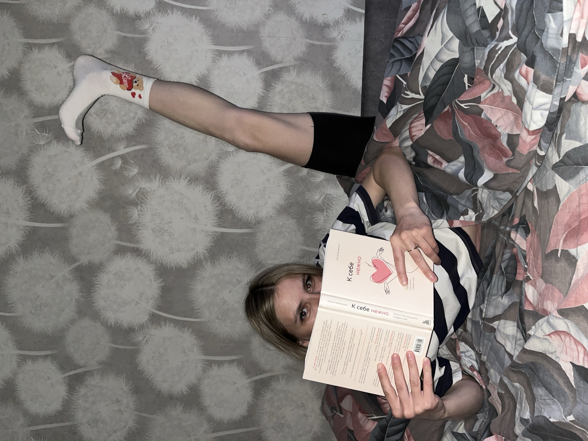 A person is lying in bed, holding a book titled "К себе нежно" ("To Self, Gently") in Russian, with their leg extended in a white sock featuring a teddy bear design. The book is open, revealing Russian text. The person is wearing a striped shirt and black shorts. The bed is covered with a patterned blanket with floral designs in shades of grey, white, pink, and brown. The background features wallpaper with a dandelion pattern. No location is discernible within the frame.