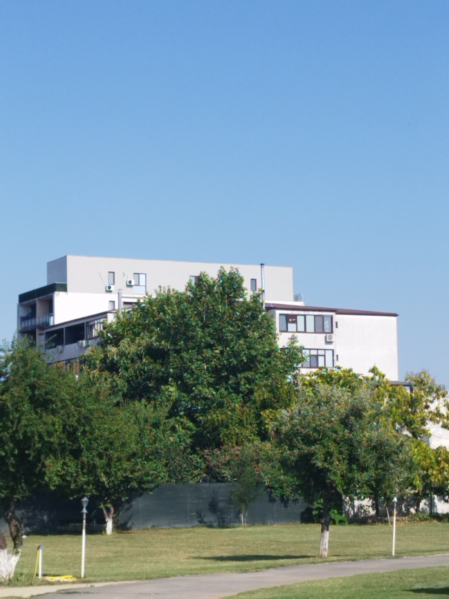 In a scene of an urban environment, a multi-story building with a modern facade stands against a clear blue sky. The building, a light gray color with white accents, features multiple windows and balconies. In front of the building, a dense canopy of lush green trees obscures a portion of its lower levels, creating a natural buffer. Beyond the trees, a dark-colored fence encloses a grassy area. The foreground displays a paved pathway and a well-maintained lawn, with a white lamppost and some yellow and white stakes visible on the grass. The overall setting appears to be a well-kept outdoor space within the city of Bucharest, Romania.