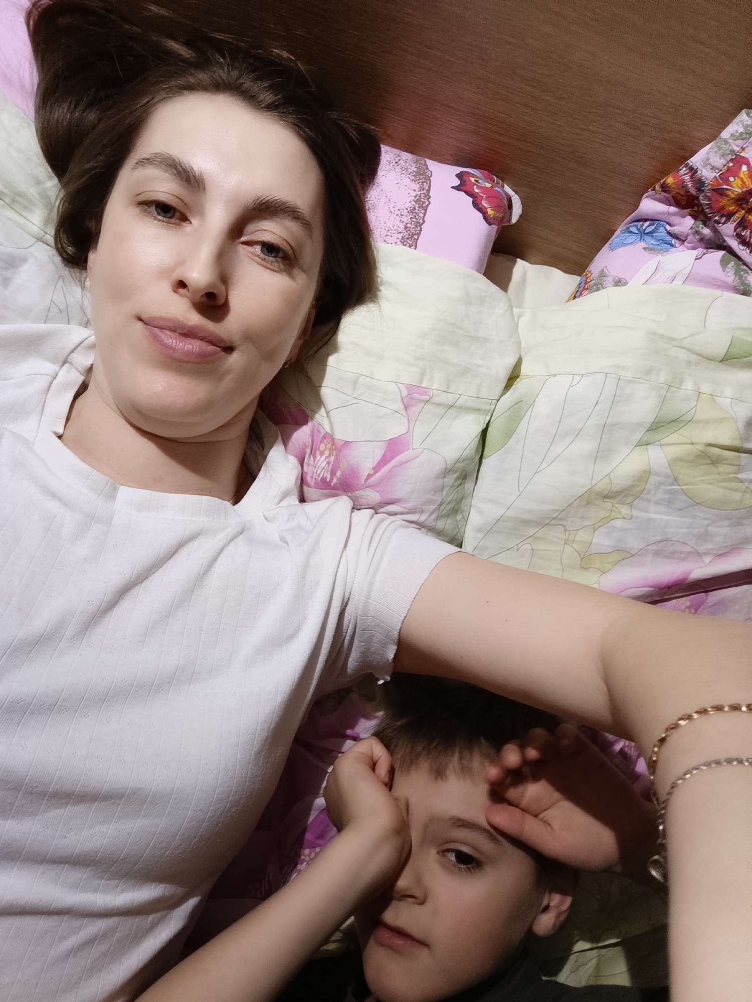 A woman and child are lying in a bed with floral patterned sheets and pillows. The woman is fair-skinned with brown hair and is wearing a white ribbed shirt. The child appears to be a young boy with short brown hair, partially obscured by his hand near his face. The woman is wearing two bracelets on her right wrist. The location is unspecified and there is no indication that it is in Chaikovsky, Russia.