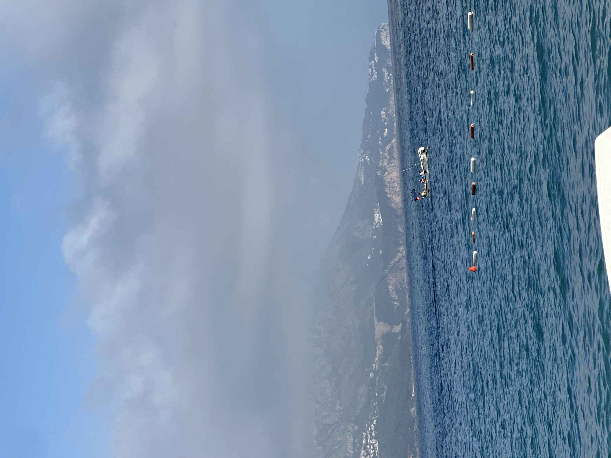 A landscape photograph shows a coastal scene, seemingly in Budva, Montenegro, featuring a body of water, a coastline with vegetation, and a partly cloudy sky. The water is a deep shade of blue, indicating depth or a clear seabed. Floating in the water is a series of white and red markers, possibly buoys, set in a line, indicating a designated zone or boundary.
Alongside the water is a shoreline covered in trees and what appear to be buildings. A thin layer of fog or cloud hangs over the trees, making the buildings difficult to discern.
The upper left portion of the photograph shows a sky with areas of blue and patches of clouds. The image is taken from an elevated point of view, perhaps from a boat or a high vantage point on the coast.
A small boat with several people is visible near the coastline and markers. The people appear to be engaged in some kind of activity near the markers.
