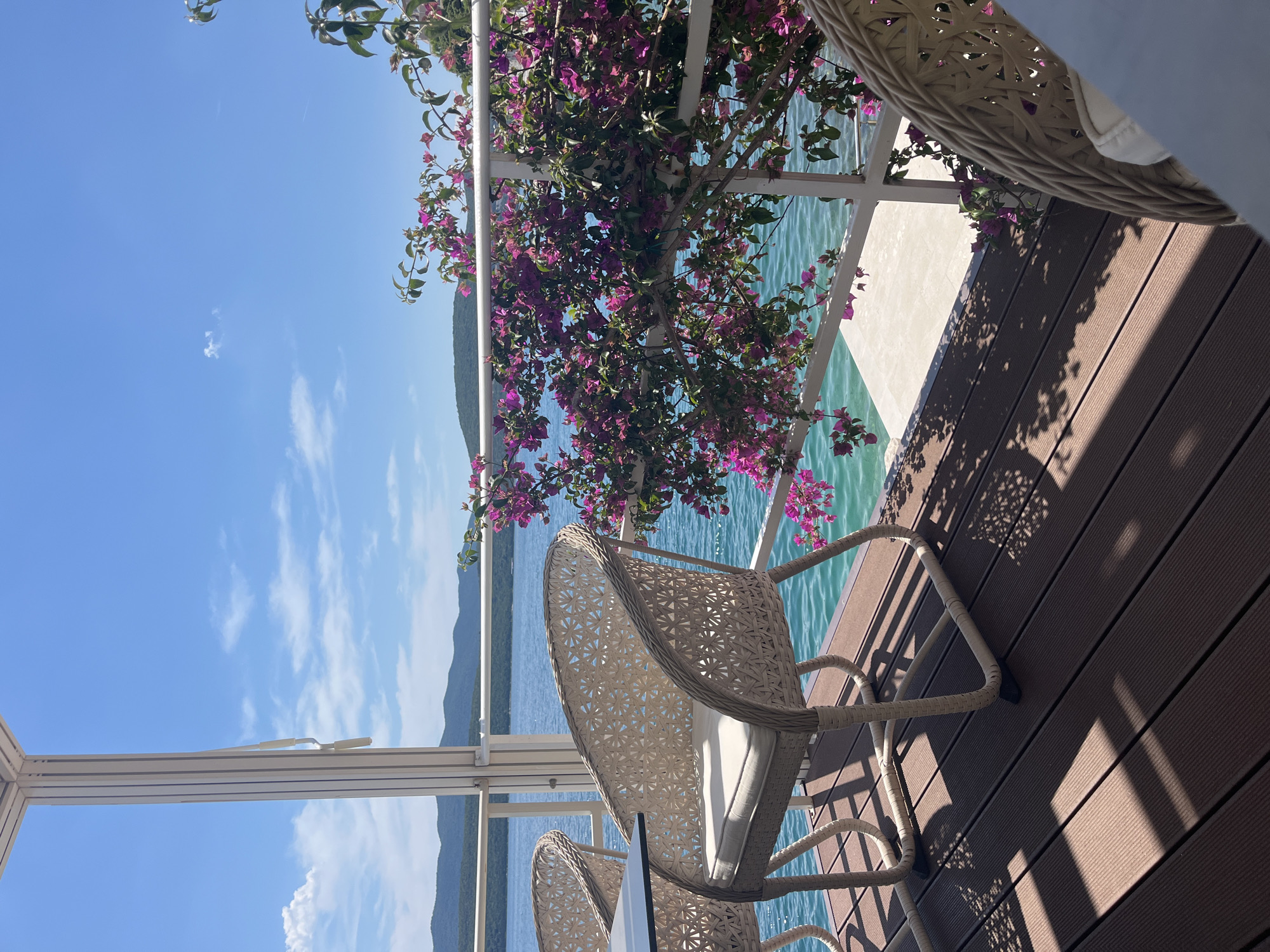 The image captures a serene outdoor scene with a focus on relaxation and the natural environment. A pair of light-colored, wicker chairs with white cushions are positioned on a wooden deck, inviting one to sit and enjoy the view. A vibrant bougainvillea plant, with its pink and purple flowers, drapes over a white railing, adding a splash of color and a sense of tranquility to the setting. The background reveals a calm body of water, possibly a sea or a large lake, with distant mountains visible on the horizon. The sky is a clear blue with scattered clouds, suggesting a bright, sunny day. The location is described as Unknown, Unknown.