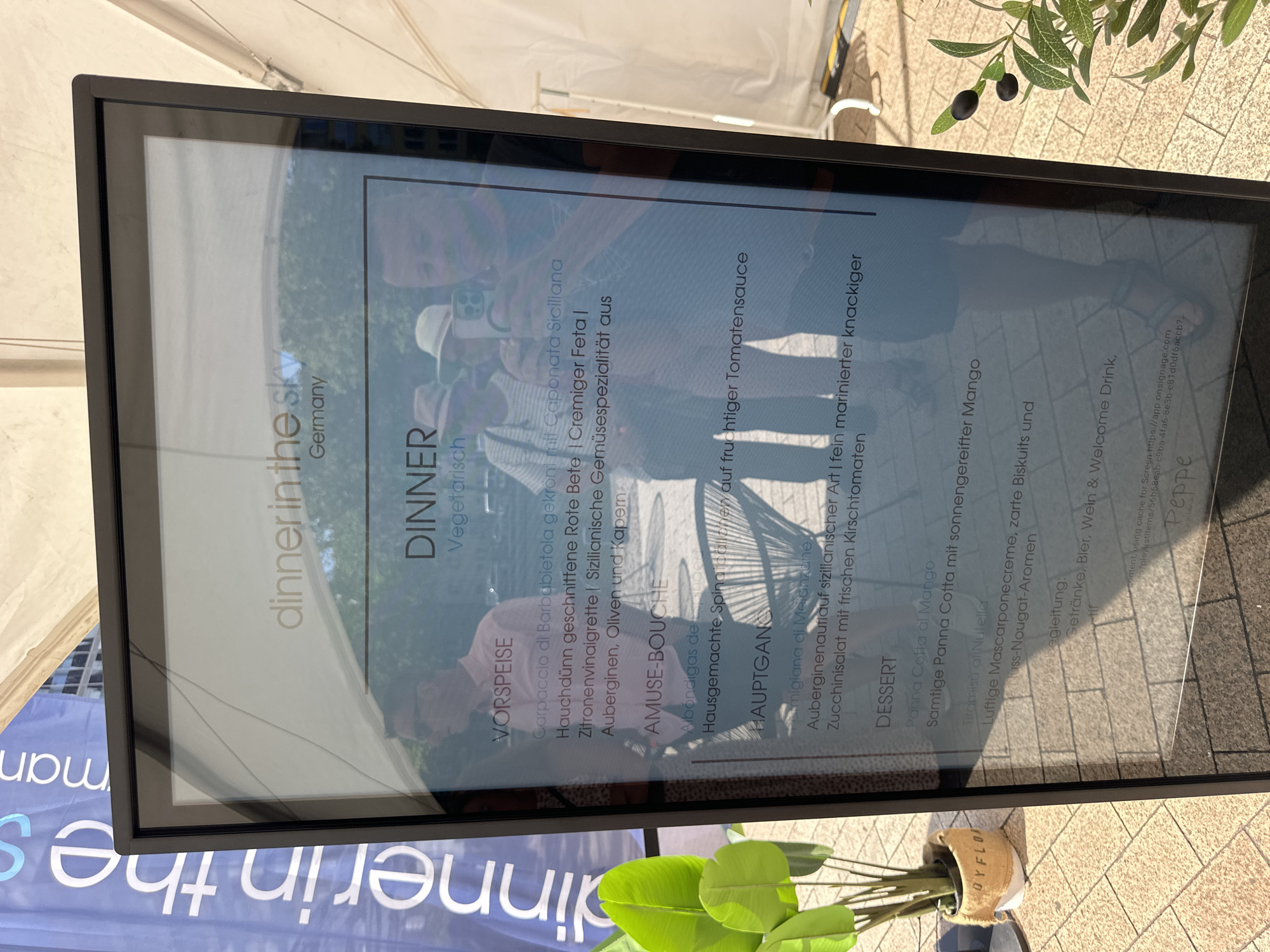 The image shows a digital menu board in front of a temporary tent structure. The menu is for "Dinner in the Sky Germany" and is located in Cologne. The menu lists "Vorspeise" (appetizer), "Amuse-Bouche", "Hauptgang" (main course), and "Dessert". The appetizer is Carpaccio di Barbabietola, the amuse-bouche is Albondigas de Espinaca, the main course is Melanzane alla Parmigiana, and the desserts are Panna Cotta al Mango and Tiramisu al Nutella. The menu also mentions a welcome drink of beer, wine, and soft drinks. In the background, a person is holding up a phone to take a picture of the menu. There is also a potted plant with green leaves and a few olives on a branch to the right of the menu board. The ground is paved with light-colored tiles.