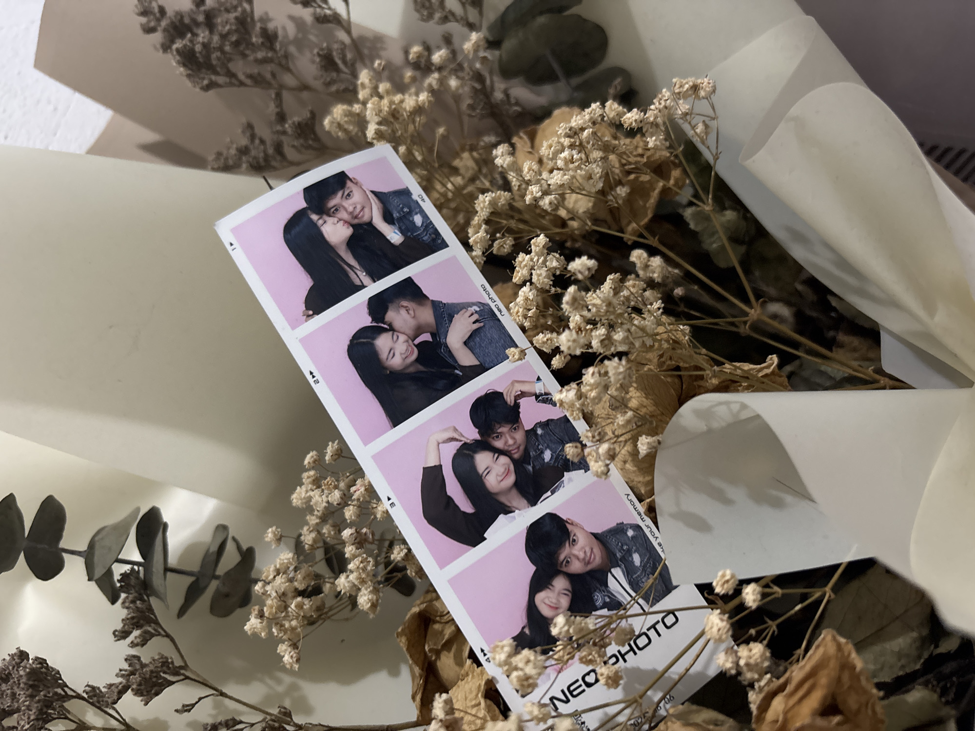 The image displays a bouquet of dried flowers with a pink photo booth strip featuring four portraits of two young couples. The photo strip is positioned diagonally across the bouquet. In the first portrait, a young woman leans in to kiss a young man on the cheek. The second portrait shows the same couple, with the woman kissing the man's cheek. The third portrait features a different young woman and man, with the woman forming a heart shape with her hands over the man's head. The fourth portrait shows a young woman and man, with the woman kissing the man on the cheek. The photo strip has "NEO PHOTO" printed on it. The bouquet is wrapped in white paper and some brown paper. The background appears to be an indoor setting with a textured wall. No specific location in North Jakarta, Indonesia is identifiable.