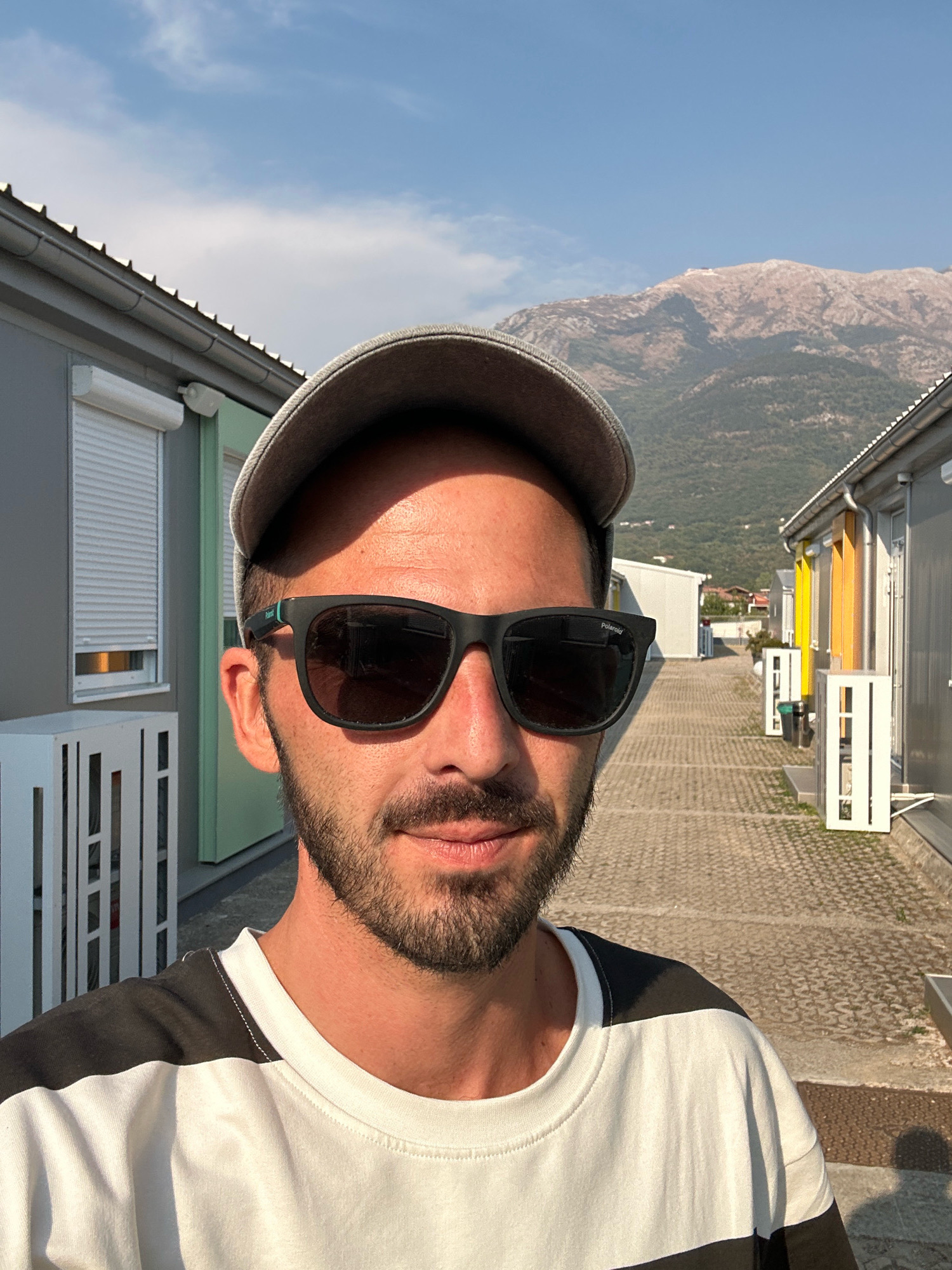 A man wearing a gray baseball cap and black sunglasses is pictured in a selfie. He has a beard and is wearing a black and white striped t-shirt. He is standing outdoors in what appears to be a residential or temporary housing area with rows of modular buildings. In the background, a large, rocky mountain range is visible under a clear blue sky. The scene is set in the city of Radanovići, Montenegro. The pavement is paved with interlocking concrete blocks, and there are white railings and decorative white structures along the path. Some of the buildings have roll-down shutters on their windows.