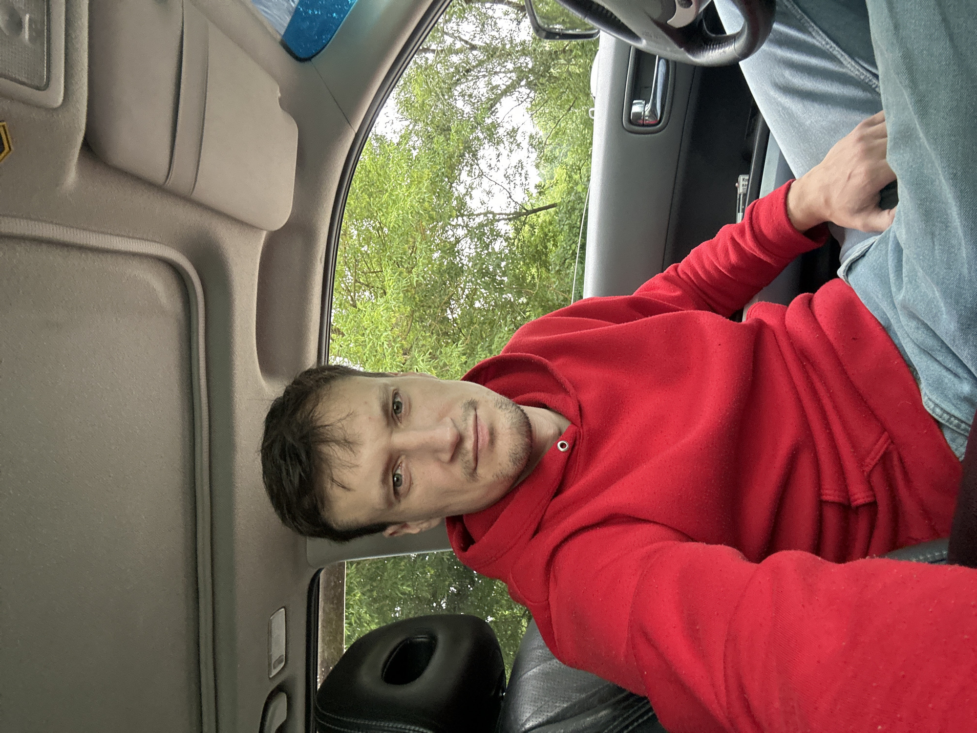 A man with short brown hair and stubble is seated in the driver's seat of a car, looking at the camera. He is wearing a red hooded sweatshirt and light blue jeans. The interior of the car is visible, including the car's ceiling, a sun visor, a black headrest, and the steering wheel. Outside the car, green trees are visible through the windows, suggesting an outdoor, possibly rural, setting. The man's left arm is resting on his lap, and his right hand is near the steering wheel. The image is taken from a slightly low angle, looking up at the man.