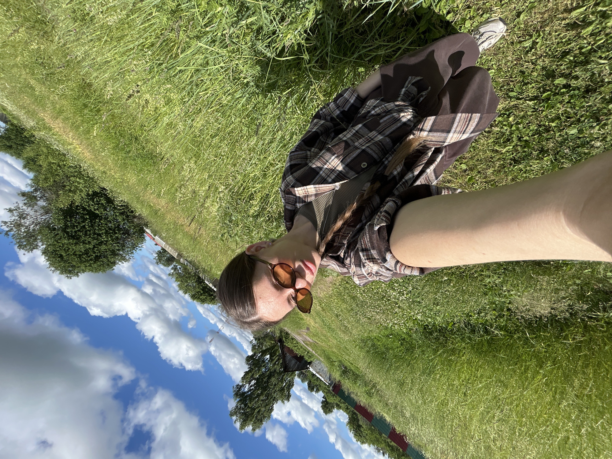 A young woman is taking a selfie outdoors in a grassy field. She is wearing sunglasses, a brown and white plaid shirt over a green t-shirt, and brown pants. Her long brown hair is braided and hangs over her shoulder. The field is covered in tall green grass, with some small white flowers visible. In the background, there are trees and a rustic wooden building. A fence with red and green sections runs along the left side of the field. The sky is blue with fluffy white clouds. The image appears to have been taken on a sunny day. The location is in the city of Malaya Borshchevka, Russia.