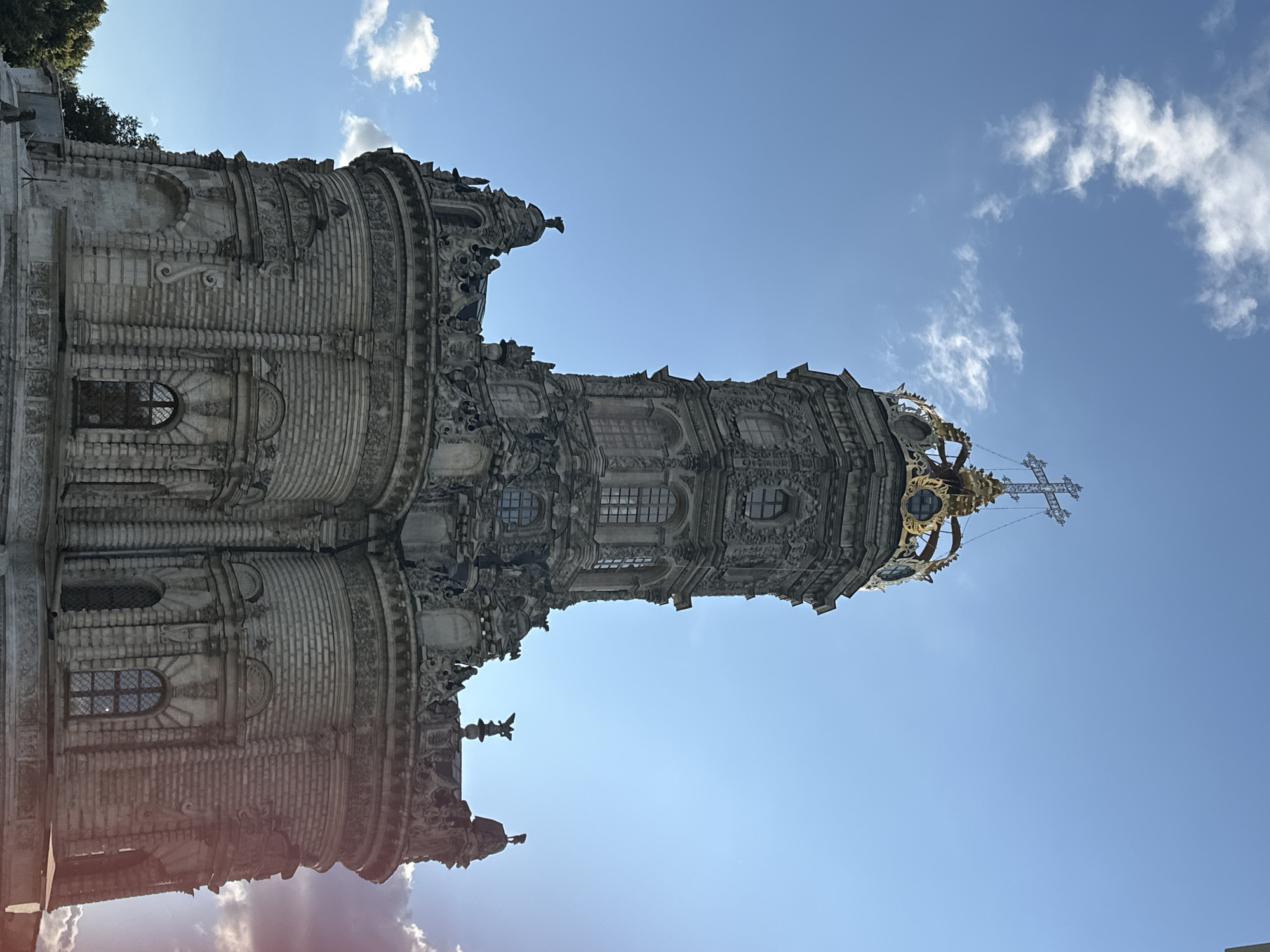 The image captures a low-angle, full shot of the Church of the Sign of the Most Holy Theotokos in Dubrovitsy, Russia, against a bright blue sky with scattered white clouds. The church's distinctive architecture, characterized by its ornate Baroque style, is prominently displayed, featuring multiple tiers, decorative carvings, and a large, gilded crown-like structure topped with a cross. Sunlight illuminates the church, casting some areas in shadow and creating lens flare on the right side of the image. The surrounding environment is only partially visible, with hints of trees on the left and the edge of a paved area in the foreground, suggesting an outdoor setting. There are no people or animals visible in the frame.
