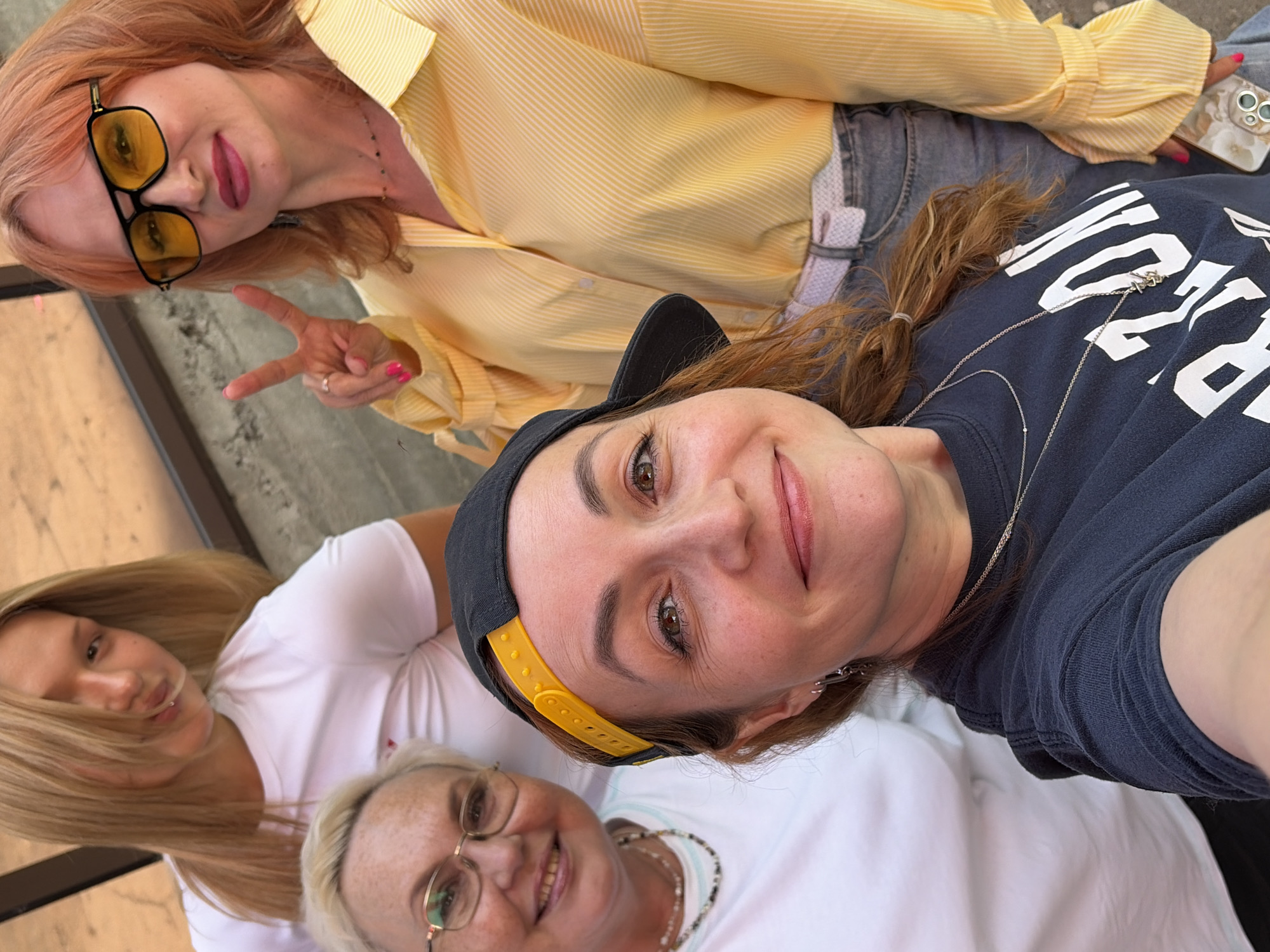 A rotated eye-level, medium shot shows four people, one wearing a black baseball cap with a yellow adjustable strap, posing for a selfie. The person taking the selfie is wearing a dark-blue t-shirt, and has light skin, brown hair, and brown eyes. Two women are smiling below, and one blonde woman is off to the side. The fourth woman is in the upper right, wearing a yellow striped button down shirt, sunglasses and holding up a peace sign. The surrounding environment isn't clearly visible but there are gray elements indicating this might be a concrete sidewalk. The specific location is unknown.