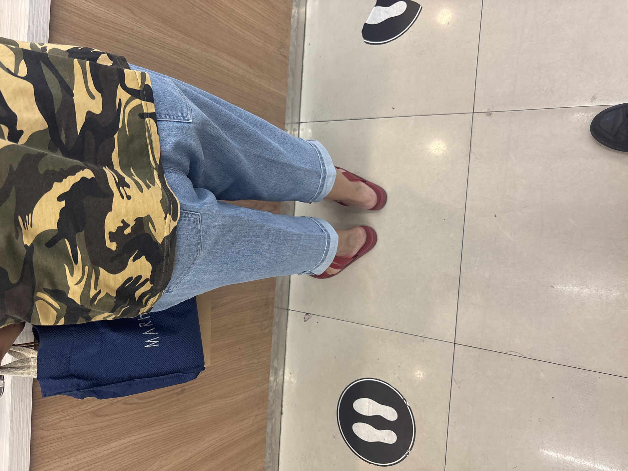 In North Jakarta, Indonesia, an individual stands on a tiled floor, facing away from the camera. They are wearing a camouflage t-shirt and light blue denim jeans cuffed at the ankles. Red flip-flops are on their feet. To their left, a navy blue tote bag with a textured handle hangs from their shoulder, displaying the white text "MARH" and part of a logo. On the floor, a black circular decal with a white footprint graphic is visible to the person's left, indicating a designated standing spot. Another similar decal is partially visible to the right. The background consists of a wooden wall.