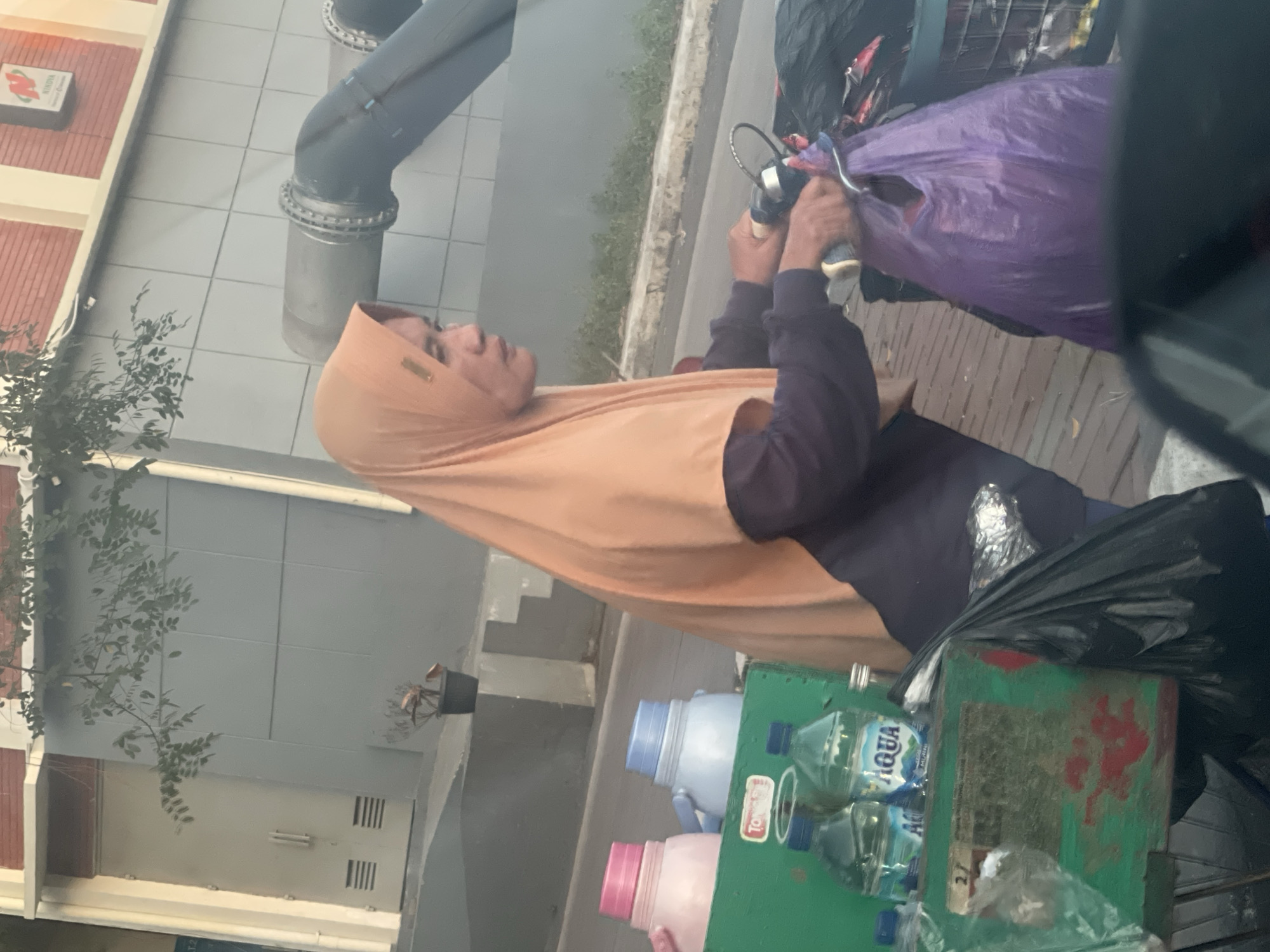 An individual wearing an orange hijab and a dark long-sleeved shirt is pushing a cart filled with items, including water bottles, plastic jugs, and trash bags. The cart is green and appears to be made of wood and metal. The individual is holding onto the handlebars of what appears to be a bicycle or tricycle attached to the cart. In the background, there is a building with a gray facade and a red brick section, along with large pipes. The scene is set on a paved walkway next to a road. The lighting suggests it is daytime.