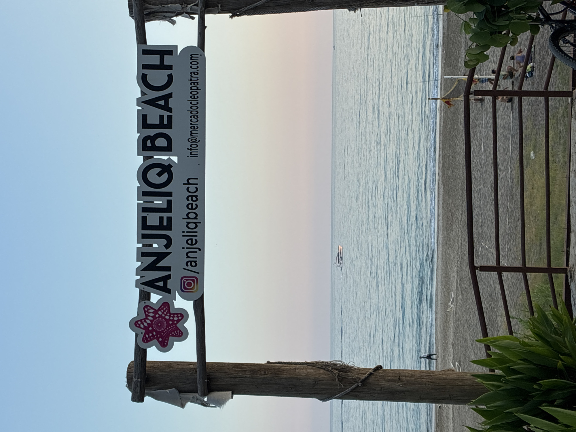 This image depicts a scenic view of Anjelik Beach in Alanya, Turkey, as indicated by the signage visible in the frame. The scene is captured at what appears to be either sunrise or sunset, with a soft gradient of pastel colors in the sky.
In the foreground, a wooden archway frames the view of the beach and the sea. One of the wooden posts has a weathered texture and is adorned with a piece of frayed white fabric and a length of rope. The archway supports a sign that reads "ANJELIQ BEACH" in bold black lettering. Below this, an Instagram logo and the handle "/anjeliqbeach" are displayed, along with an email address: "info@mercadocleopatra.com". A decorative pink starfish-like emblem is also attached to the sign.
The beach itself is sandy, with a gentle slope towards the sea. A person is seen in the distance performing what looks like a handstand on the sand, facing the water. Further down the beach, a group of people are gathered near a volleyball net, suggesting recreational activities.
The sea is calm and appears to be a light blue, reflecting the soft hues of the sky. A small white boat is visible in the distance, moving across the water. Lush green foliage is present in the lower left and right corners of the image, hinting at the surrounding vegetation. A metal railing runs horizontally across the lower part of the frame, separating the viewpoint from the beach area. A bicycle wheel is also visible in the bottom right corner, partially obscured by the foliage. The overall atmosphere is tranquil and inviting.