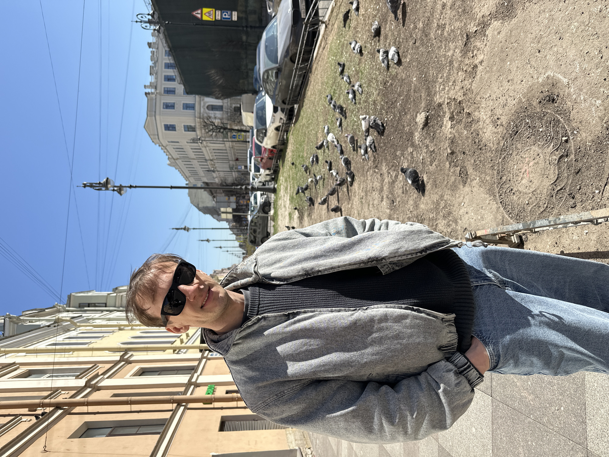 A man stands on a sunny city sidewalk in Saint Petersburg, Russia, looking towards the camera with a slight smile. He is wearing dark sunglasses, a light gray acid-wash denim jacket, a dark ribbed sweater, and blue jeans, with his hands in his pockets. The sky above is a clear, bright blue, indicating sunny weather.
To the man's right, an unpaved strip of ground with sparse grass hosts a large flock of pigeons. Beyond this, several cars are parked along the street. Multi-story buildings, typical of Saint Petersburg's classical architecture, line both sides of the street. The building directly behind the man has a light, possibly yellow or beige facade, while the one across the street is a lighter, ornate building. Numerous utility poles with overhead wires are visible against the sky.
Strong sunlight casts distinct shadows, suggesting it is daytime. A decorative manhole cover is visible on the ground amidst the dirt where the pigeons are. On the building to the man's left, a partially obscured green sign with white text appears to read "Новая Аптека" (New Pharmacy). In the upper right background, traffic signs, including a yellow diamond (priority road) and a blue 'P' (parking), are visible. The scene depicts a casual moment in a bustling urban environment.