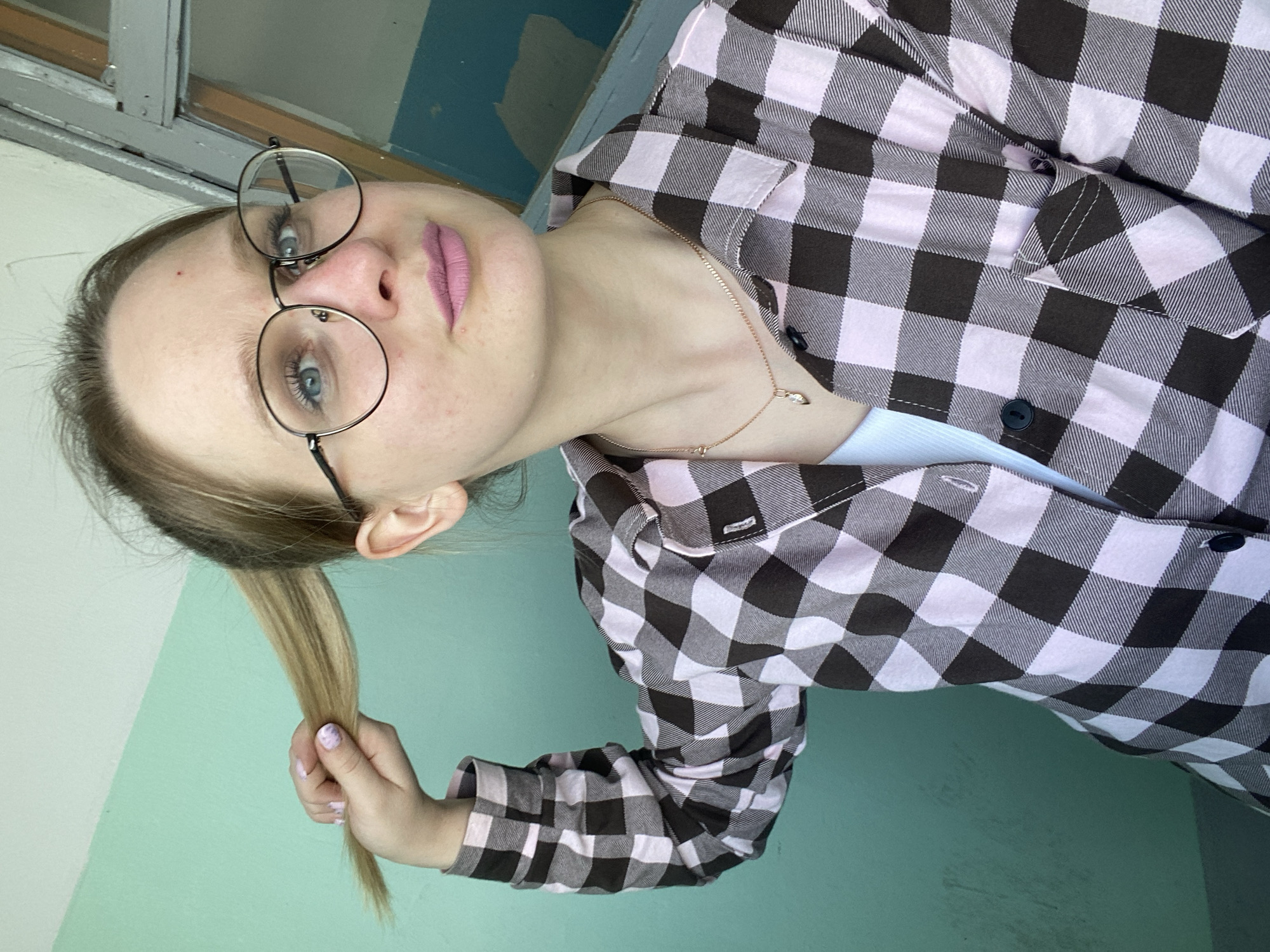 A person is holding a section of their blonde hair in their right hand, with nails painted a light purple. They are wearing round glasses and a pink and black checkered shirt over a light blue top, with a gold necklace visible. The background consists of a turquoise wall and a white-framed window. The location is Unknown, Unknown.