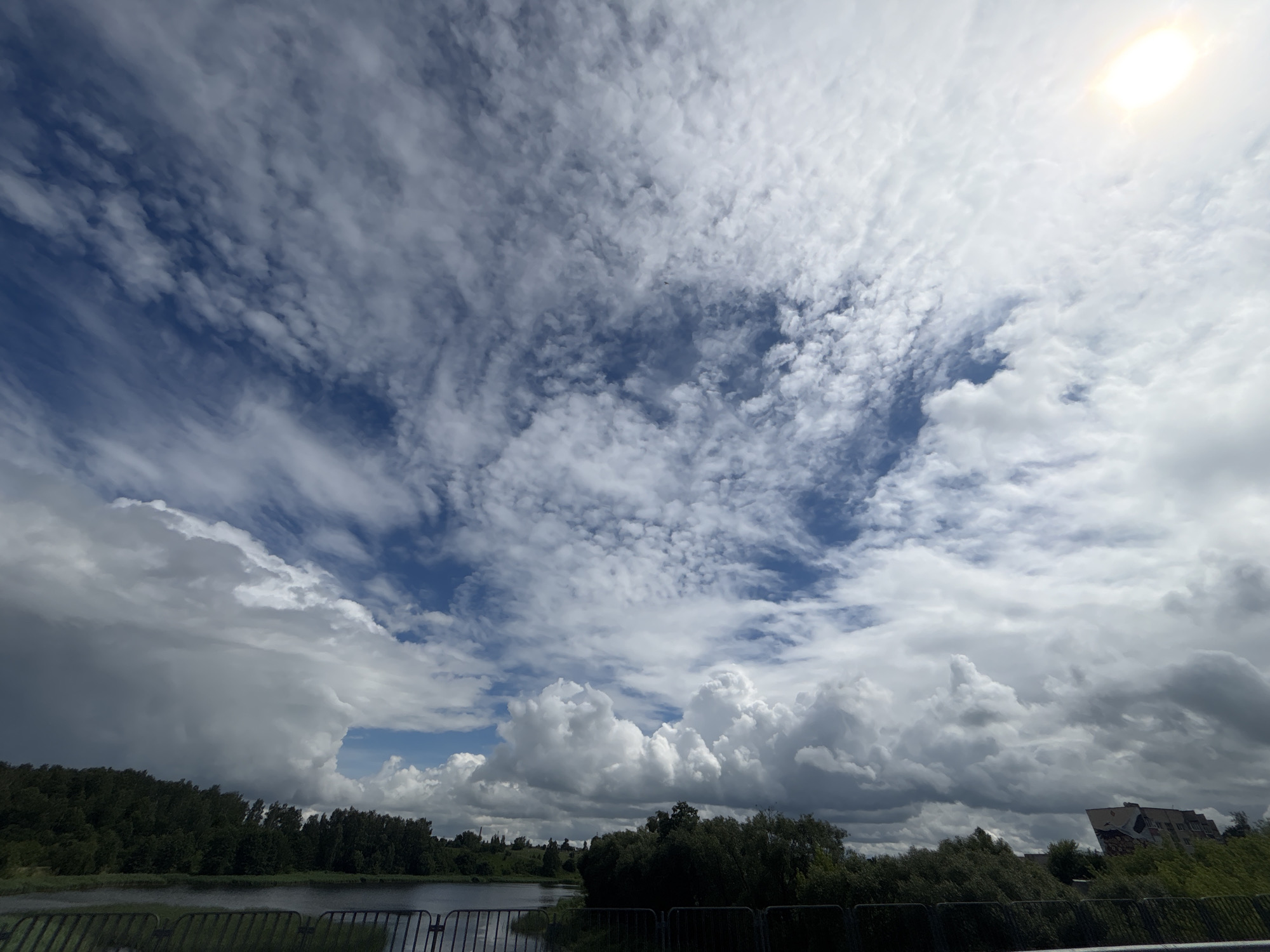 This is an eye-level, wide-angle shot capturing a dynamic sky over a rural landscape in Капыльскі сельскі Савет, Belarus. The scene depicts a large body of water in the foreground, separated from the viewer by a dark metal fence. Beyond the water, a dense line of trees forms a dark green border separating the water from the sky.
The sky is the dominant subject, filled with a variety of clouds. High in the sky, wispy cirrus clouds streak across a bright blue expanse. Lower down, cumulus clouds form larger, puffy masses, with some appearing dark and stormy, suggesting impending rain. The sun is visible in the upper right corner, a bright, blurred orb peeking through the clouds, indicating it's daytime.
There are no people or distinct objects in the scene, besides the fence and the distant trees. The overall mood is one of natural grandeur, with the dramatic sky creating a sense of awe and perhaps anticipation of changing weather. The location cues, such as the specific cloud formations and the type of vegetation, are consistent with a temperate climate. There is no visible text in the image.