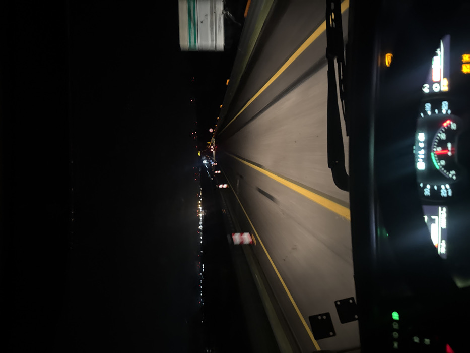 The image captures a nighttime scene from the driver's perspective inside a vehicle on a multi-lane highway near Cerignola, Italy. The roadway is illuminated by the vehicle's headlights, revealing asphalt with prominent double solid yellow lane markings. Distant artificial lights are visible on the horizon, indicating a populated area or industrial complex.
On the left shoulder, a white and red striped traffic bollard is present, suggesting roadwork or a temporary lane boundary. Vehicle lights, including red taillights and white headlights, are discernible in the distance, indicating other vehicles on the roadway. A rectangular green and white road sign is partially visible on the right side of the frame.
The lower section of the image displays the vehicle's dashboard, featuring illuminated instruments. A speedometer with a red needle, a tachometer with a green arc, and several active indicator lights are visible, including a green light on the left and an amber light on the upper right. A black windshield wiper rests at the base of the windshield. The overall environment is dark, with artificial light sources providing the primary illumination.