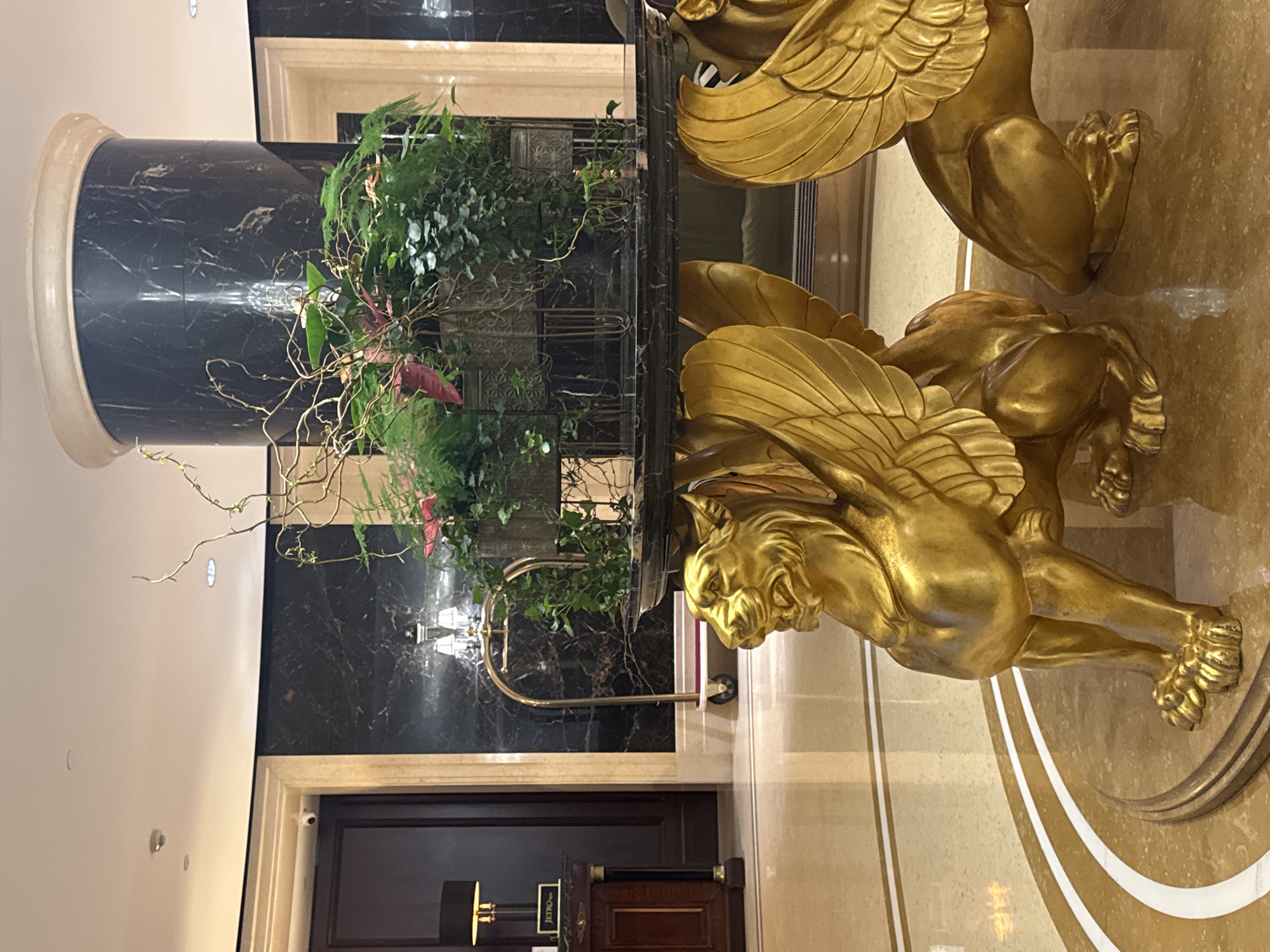 The image depicts the lobby of a luxurious establishment, likely a hotel or upscale building, in Unknown, Unknown. A black marble column with a circular, cream-colored top and base is positioned on the left side of the frame, decorated with a large green plant arrangement. The column appears to support a glass table. To the right of the column is a large, gilded sculpture of winged lion-like creatures, apparently holding up the table. The floor is marble with gold and white accents. In the background, a dark wooden door is visible, along with luggage carts. The overall ambiance suggests opulence and sophistication.