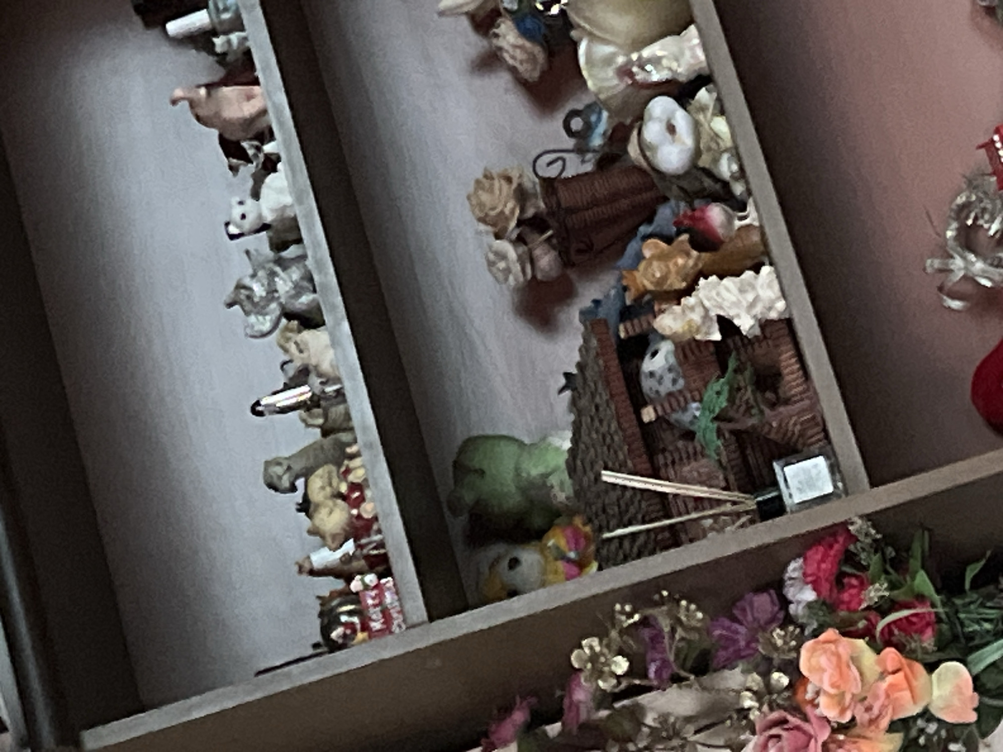 The image shows a section of a shelf or cabinet, densely packed with various small objects and figurines. On the left side, numerous animal figurines, including what appear to be pigs and dogs, are arranged vertically. In the middle section, there are decorative items, including floral arrangements, ribbons, and a small bird figurine perched on a miniature house. The bottom shelf is filled with artificial flowers in a range of colors. There is a white, square object next to wooden sticks. The overall impression is one of cluttered storage or display. The city and state are unknown.