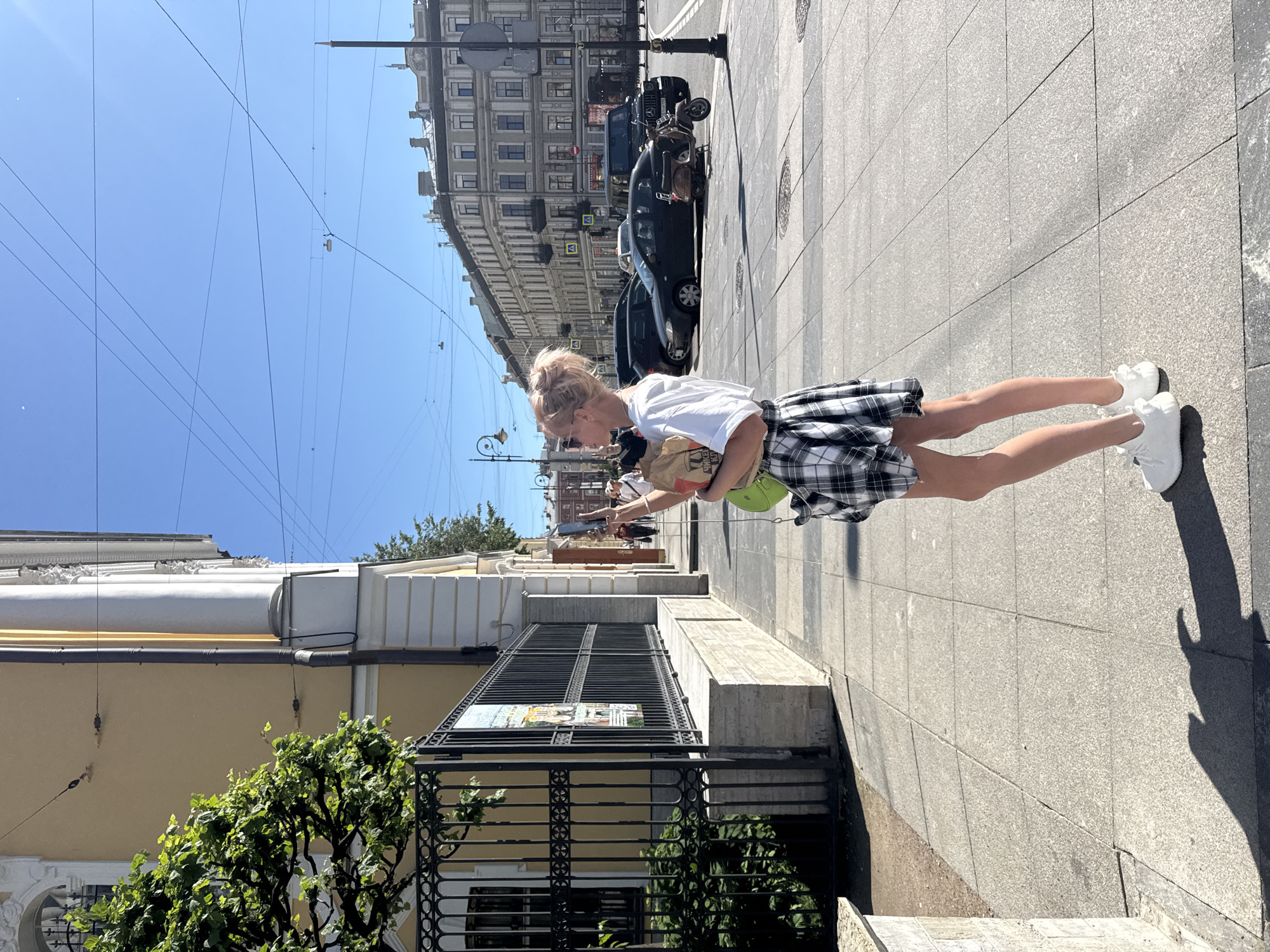 A young woman with blonde hair styled in a bun stands on a sidewalk in a city, likely Saint Petersburg, Russia, given the architectural style of the buildings and the presence of overhead wires. She is wearing a white t-shirt, a black and white plaid skirt, and white sneakers. A green crossbody bag is visible. She appears to be holding a paper bag in her right hand, and her left hand is extended, possibly interacting with something or someone not fully in view.
The setting is a sunny day, indicated by the clear blue sky and the distinct shadows cast on the pavement. The sidewalk is paved with large grey tiles, and a black metal fence with decorative elements runs along the side of a building. Behind the fence, a yellow building with ornate white trim is visible, along with lush green foliage. Across the street, there are more buildings with classic European architecture, and several dark-colored cars and a scooter are parked.
The overall mood seems relaxed and ordinary, as if the woman is out for a stroll on a pleasant day. The clear sky suggests it is daytime, likely mid-morning or afternoon. There is no visible text in the image.