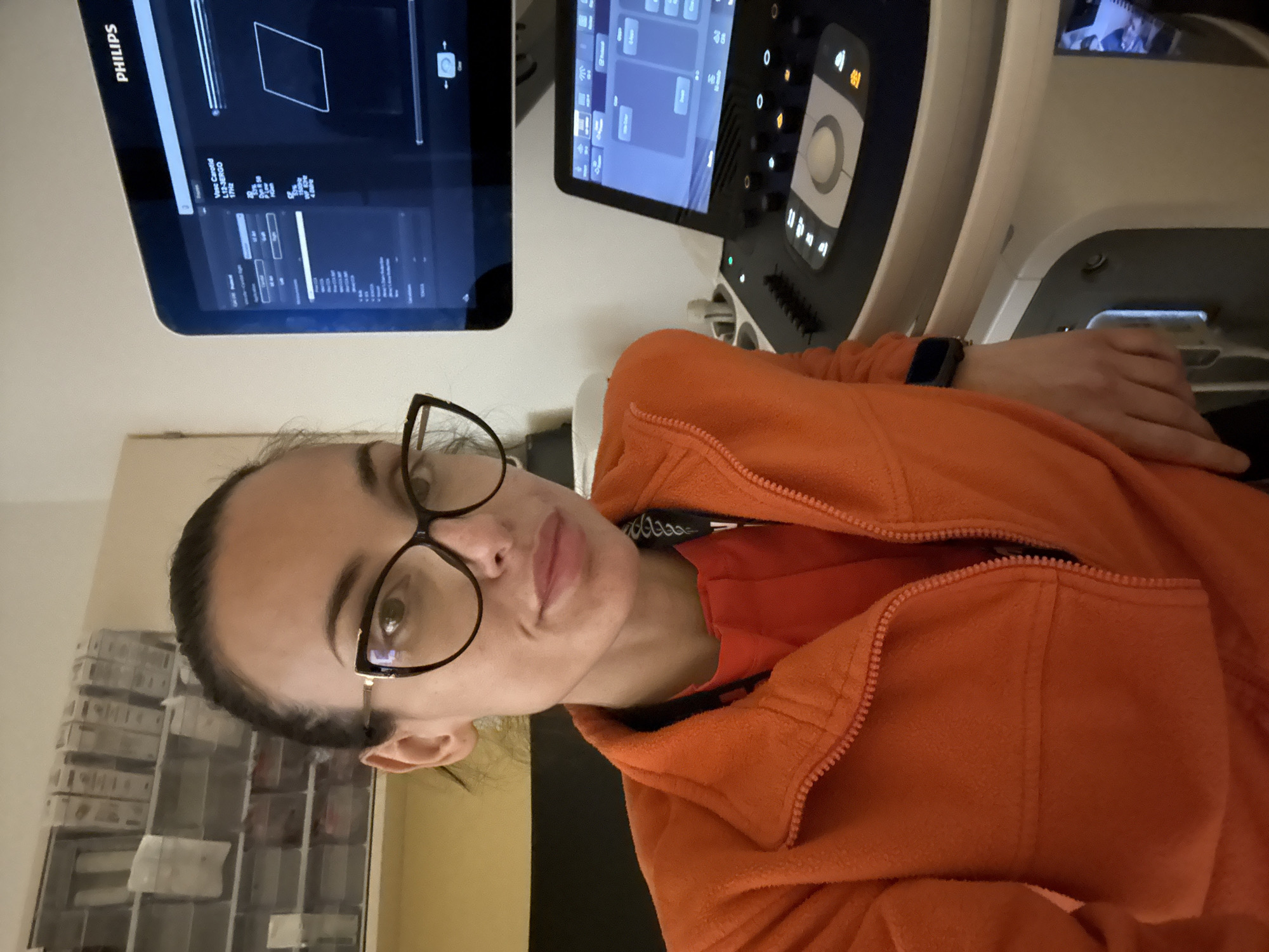 A woman with dark hair pulled back and wearing glasses is positioned in front of a Philips ultrasound machine. The machine features two screens; one displays a schematic diagram of a rectangle, while the other displays control options. She is wearing an orange jacket over a red shirt. In the background, a wall with shelving holding boxes is visible. The location within the city of Pleven, Bulgaria, is not discernible from the image.