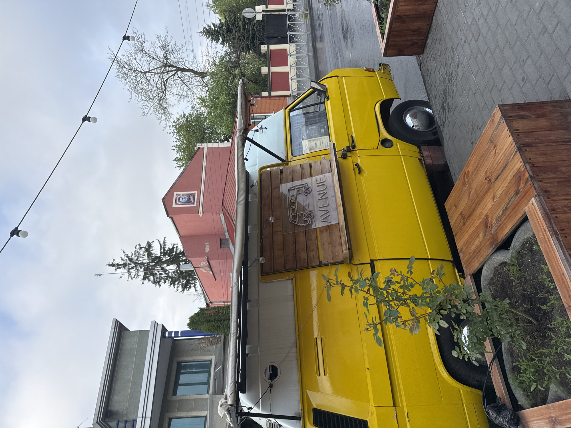 A yellow Volkswagen van, converted into a food truck, is parked on the side of a street in Akhieli, Georgia. The van, labeled "AVENUE," has a wooden sign attached to its side and an awning extended. The surrounding environment includes buildings of varying architectural styles, trees, and wooden planters. String lights are visible overhead. The ground appears to be wet, suggesting recent rain. No people are visible.