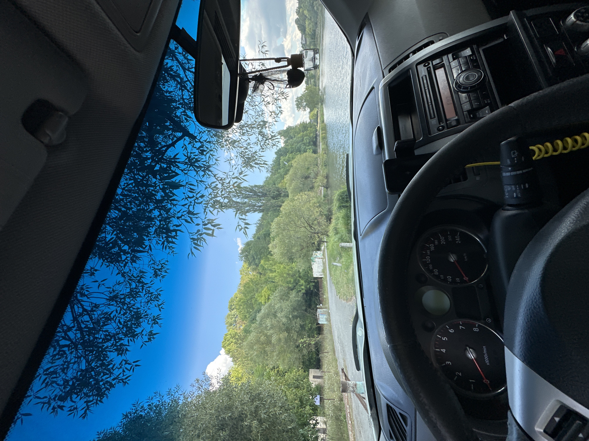 The image is taken from inside a car, looking out through the windshield. The view displays a tranquil waterscape with a body of water, likely a lake or river, surrounded by lush green trees and vegetation under a bright blue sky with scattered clouds. In the foreground, the car's dashboard is visible, including the steering wheel and instrument cluster. A rear-view mirror hangs from the top of the windshield, with an air freshener dangling from it. The scene suggests a peaceful outdoor setting, potentially a recreational area or a quiet spot by the water. The location is identified as Makiv, Ukraine, which is a village in the Cherkasy Oblast of Ukraine.