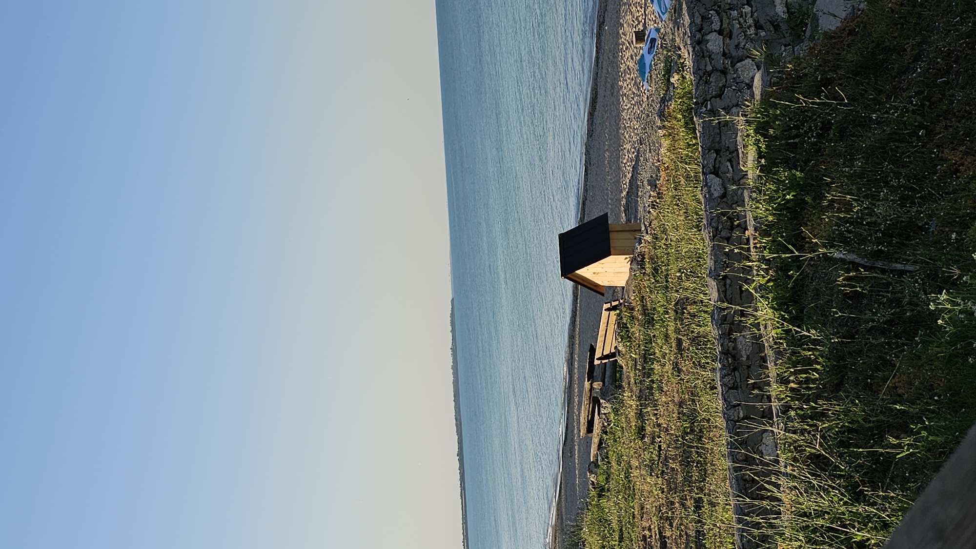 The image depicts a tranquil scene by the sea, likely in Shabla, Bulgaria. The dominant elements are the vast, calm blue sea stretching to the horizon, a sandy beach, and a grassy embankment with a small wooden structure.
The small wooden structure resembles a shelter or a rustic playhouse, complete with a dark, angled roof. In front of it, a wooden bench is visible, suggesting a place for rest or observation. The beach is made of pebbles and sand, with a few blue items scattered on it, possibly beach chairs or mats.
The grassy embankment is overgrown with green grass and some wildflowers, creating a natural border between the beach and a rocky, possibly man-made wall. The overall setting is peaceful and natural, typical of a coastal environment.
The time of day appears to be late afternoon or early evening, as indicated by the warm, soft light casting gentle shadows. The sky is clear and a pale blue, transitioning to a lighter hue towards the horizon, suggesting fair weather. The clear water and the calm atmosphere evoke a sense of serenity and relaxation. There is no visible text in the image.