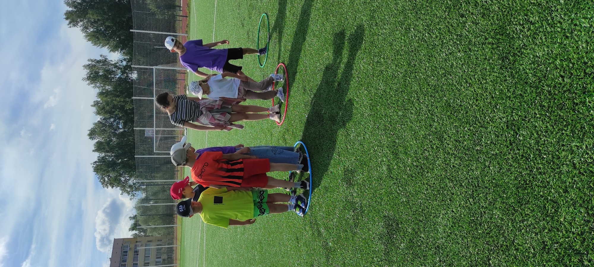 In Noviy, Russia, on a bright, sunny day, a group of children and one adult are gathered on a bright green, artificial turf sports field. They are participating in an outdoor activity involving hula hoops. Six children, dressed in casual athletic wear, are arranged around three different colored hula hoops: one blue, one red, and one green. The children appear to be of varying ages, from approximately 6 to 12 years old. One child, wearing a neon yellow t-shirt and green shorts, stands inside the blue hula hoop. To his right, another child in an orange and black striped t-shirt and red shorts is also within the blue hula hoop. A third child, in a purple t-shirt and denim jeans, stands between the blue and red hula hoops. Inside the red hula hoop, a young girl in a white t-shirt and light brown pants is positioned, and next to her, a child in a purple t-shirt and dark shorts is standing inside the green hula hoop. An adult, a woman wearing a striped t-shirt and plaid shirt tied around her waist, stands near the red hula hoop, observing the children. In the background, a chain-link fence encloses the field, beyond which are trees and a multi-story building, characteristic of an urban or suburban setting. The sky is partly cloudy with cumulus clouds.