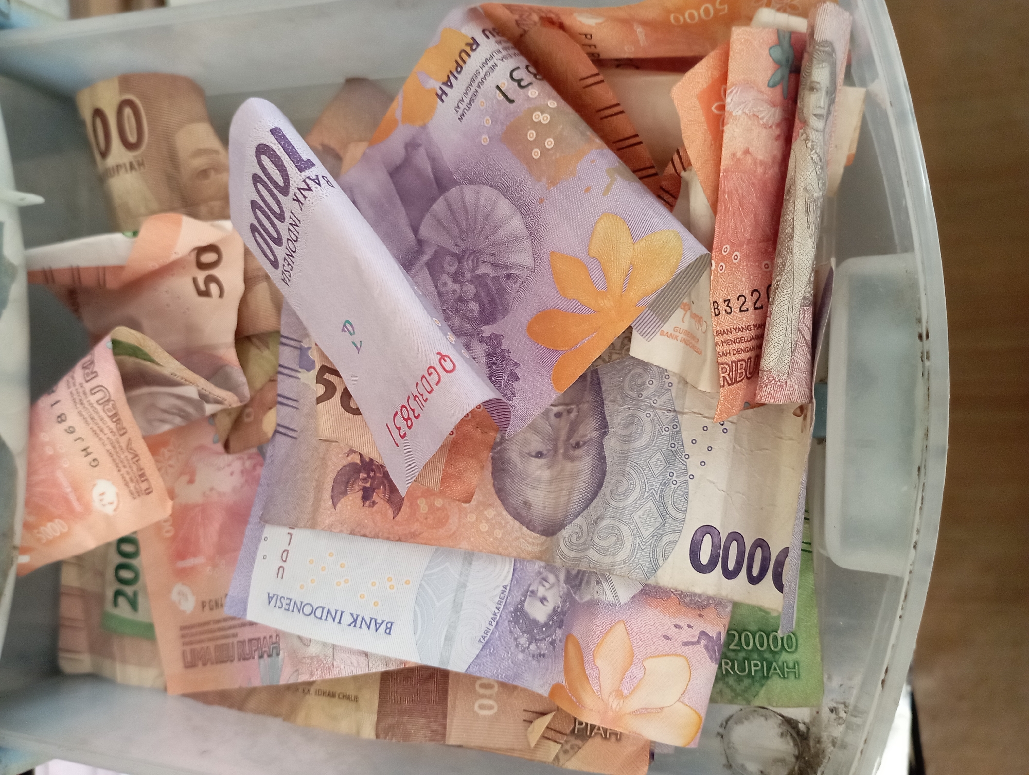 The image shows a close-up, overhead view of several Indonesian Rupiah banknotes haphazardly placed inside a light blue plastic container. The denominations visible include 200,000, 100,000, 50,000, 20,000, 10,000, and 5,000. The notes are a mix of colors, including green, orange, purple, and pink, and feature various images of people and national symbols. The container appears to be a drawer or box, with the bottom partially visible. The background suggests a domestic or office setting, with a wooden surface visible below the container. No people are present in the image, and the focus is solely on the currency and the container. The image was taken in Sembung Anyar, Indonesia.