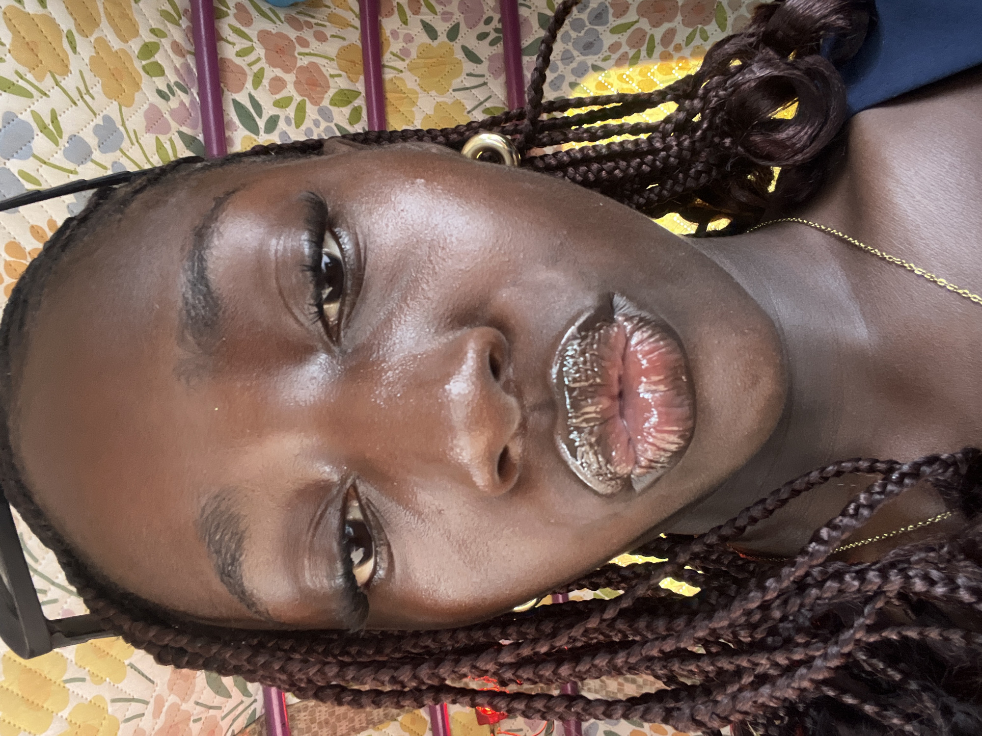A close-up photograph captures a dark-skinned woman with braided hair. She is lying on a patterned floral surface. A gold hoop earring is visible on her left ear. She is wearing a thin gold necklace. The woman is facing the camera directly with glossy lips. The background is a patterned surface, likely a bed or bedding, with floral and geometric shapes in yellow, pink, and blue. The photograph does not contain elements to determine the location in Kuchingoro, Nigeria.