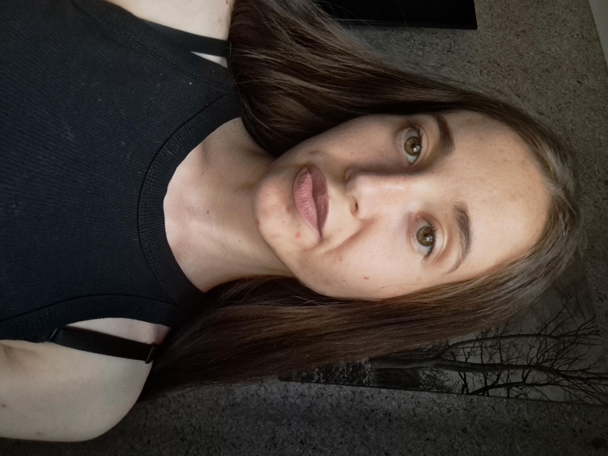 A young woman with long, brown hair is pictured from the chest up. She is wearing a black tank top. Her skin appears to have some blemishes. The background is textured with a dark pattern that resembles trees. The image was taken indoors.