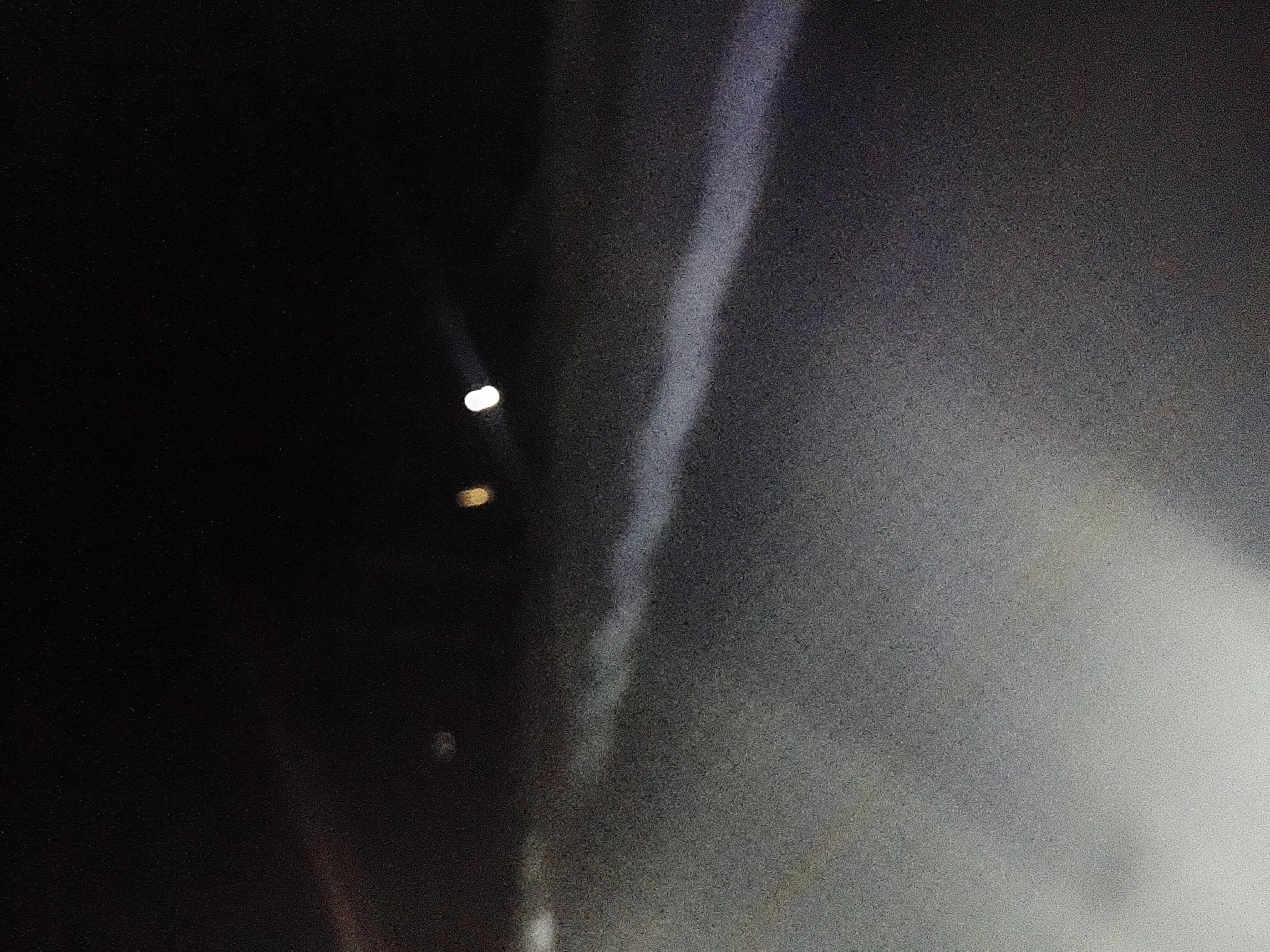 The image depicts a dimly lit outdoor scene, likely at night, with a prominent light streak extending across the frame. The streak appears to be a beam of light, possibly from a flashlight or vehicle headlights, illuminating a dusty or hazy surface. In the upper portion of the image, there are a few blurred points of light, suggesting distant light sources or reflections. The ground or surface appears to be granular and uneven. The overall atmosphere is dark and somewhat indistinct. There are no identifiable landmarks or structures that definitively place this image in Jalingo, Nigeria, or any other specific city. The image is too abstract and lacks sufficient detail for a precise location to be determined.