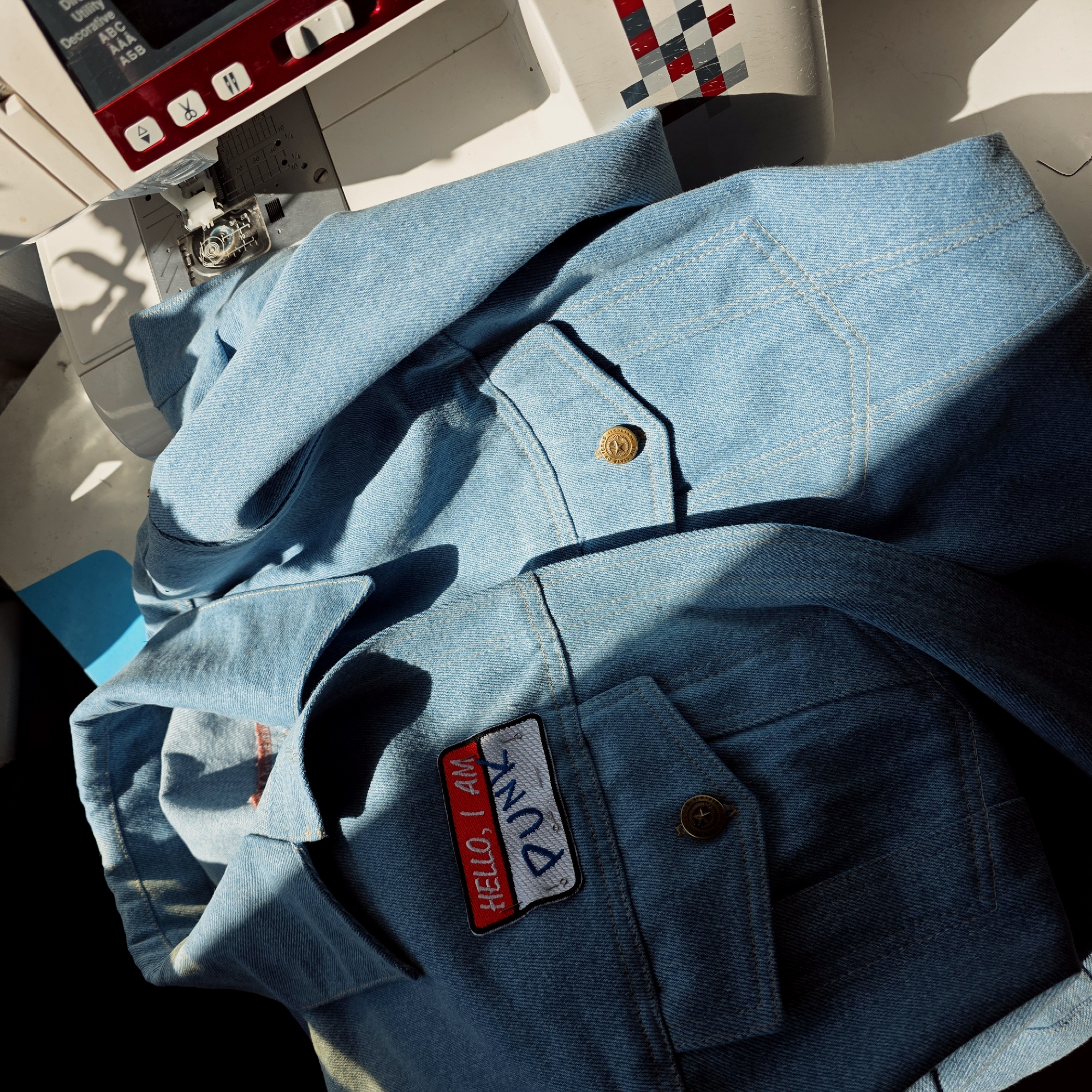 An overhead shot reveals a light blue denim garment, possibly a jacket or shirt, draped across a white and red sewing machine in an indoor setting. The scene is brightly lit by direct sunlight, casting distinct shadows and highlighting the texture of the denim fabric, along with its contrasting white stitching.
The denim piece features several notable details. A prominent rectangular embroidered patch is affixed to one of the garment's pockets or front panels, boldly displaying the text "HELLO, I AM PUNK" in red and blue letters on a white and red background. Nearby, a brass-colored button with an embossed star design is visible, and the text "TEXAS" can be discerned on its surface.
The sewing machine, partially in frame, has a modern design with a red display panel. Visible text on the machine's interface includes "Utility," "Decorative," "ABC," "AAA," and "АБВ," alongside icons such as scissors. The bobbin case, containing a spool of thread, is visible through a clear cover on the machine's bed, suggesting active use or preparation for sewing. Ruler markings like "10," "20," "30," and "40 1/4" are etched into the machine's surface. A small, bright blue object is partially visible on the left.
The overall impression is one of crafting or customisation, likely a personal project, captured during daytime. The context provided places this creative activity in Saint Petersburg, Russia.