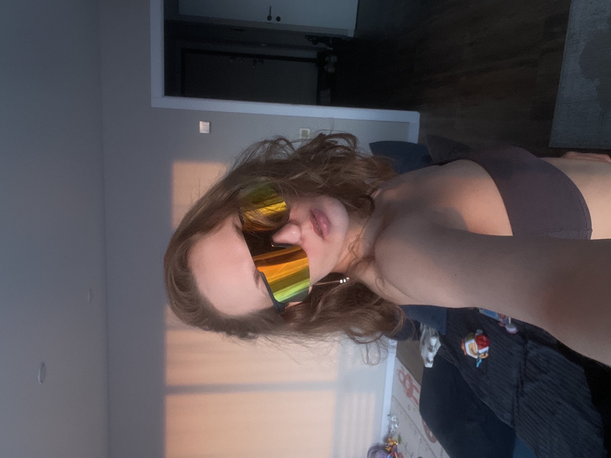 The image, taken in Подшивалово, Russia, features a young woman captured in a close-up, indoor shot. Her head is tilted to the right, with her light brown, wavy hair flowing around her face and shoulders. She wears prominent, large, wraparound sunglasses with highly reflective, iridescent lenses that display a spectrum of yellow, orange, and green hues, obscuring her eyes. Her lips are slightly parted, suggesting a relaxed or subtly pouty expression.
She is dressed in a dark-colored top, possibly a sports bra or bikini top. The background consists primarily of a plain, light grey wall. A small, square light switch or outlet cover is visible on this wall. To the right, a warm, orange-pink glow, characteristic of late afternoon or early evening sunlight, illuminates a section of the wall and her skin. In the upper right corner, a dark opening or furniture piece is visible. Below her right arm, a dark, patterned fabric with a small, colorful emblem, possibly a cartoon character like a clownfish, is faintly seen. The overall scene conveys a casual and relaxed atmosphere. There is no visible text in the image.