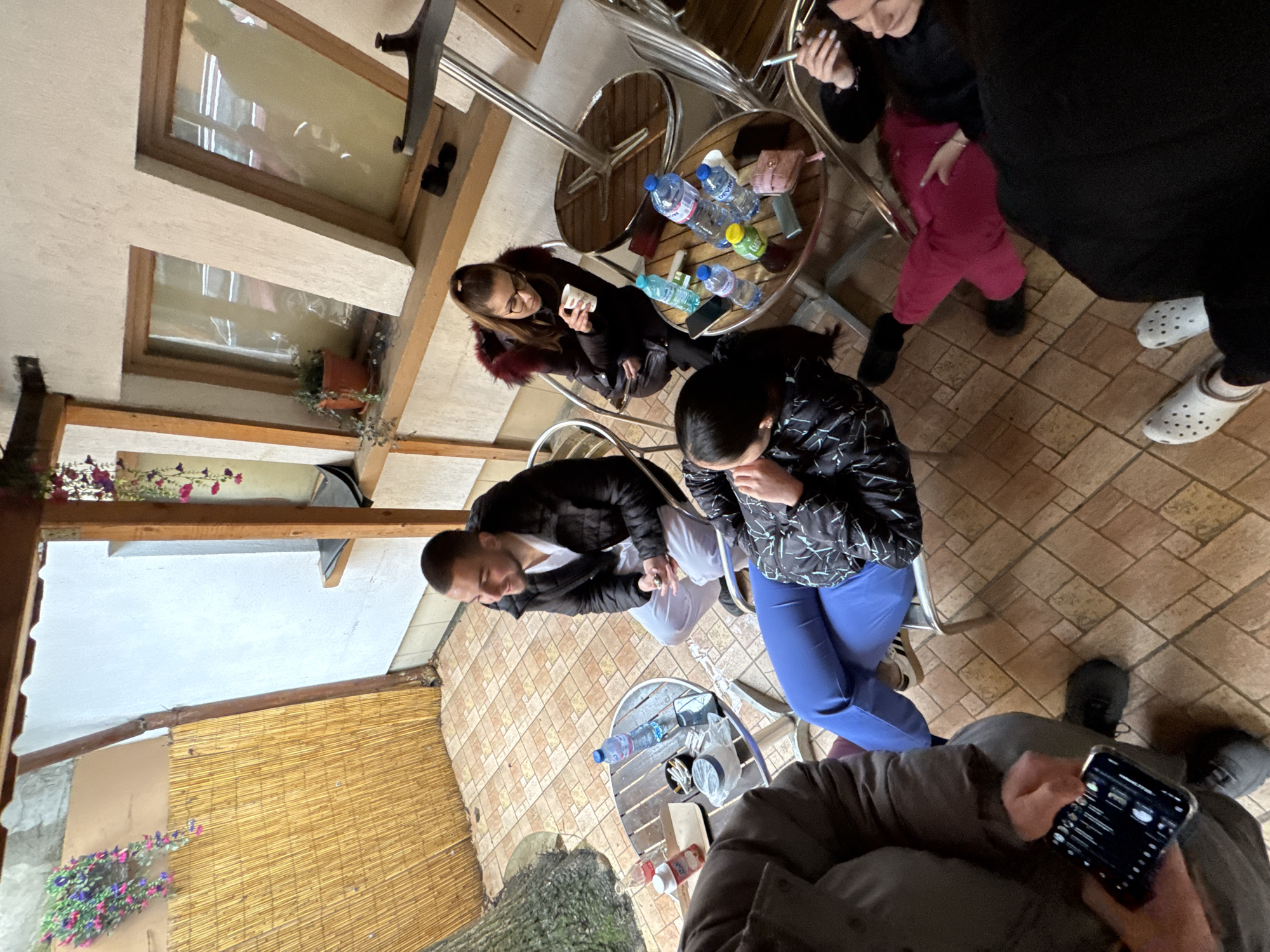 The scene depicts an outdoor seating area, likely a cafe or restaurant, in Pleven, Bulgaria. Several people are seated around small, round metal tables. The flooring is tiled in a brick-like pattern. One person is holding a smartphone, possibly taking a photograph. Tables are adorned with water bottles and other personal items. The location features a combination of open-air space and some overhead structure, as indicated by a window and a partially visible ceiling. A woven bamboo screen is visible on the left side of the image, adding to the outdoor ambiance.