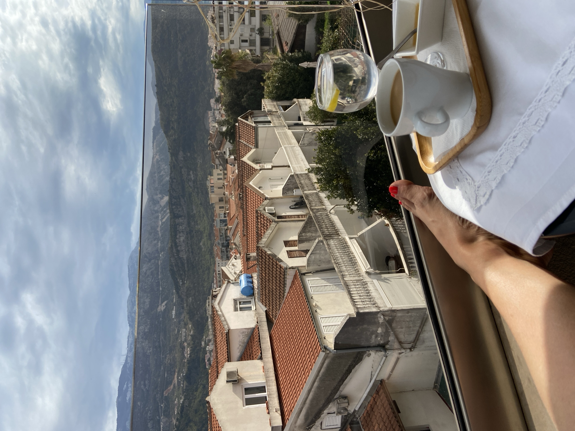 A view from a window in Budva, Montenegro. A white porcelain cup and a glass of water sit on a small wooden tray draped with a white linen cloth. A leg is visible. Outside, there are red-tiled roofs of buildings, interspersed with trees and mountains in the distance. The sky is overcast.