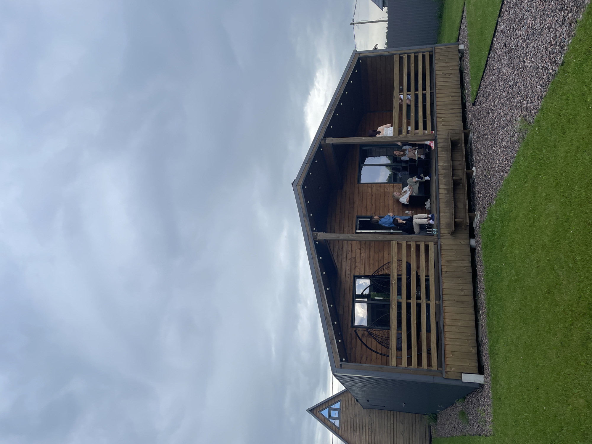 In Novo-Tuutari, Russia, a modern, wooden A-frame cabin with dark grey metal siding is situated in a grassy, outdoor setting under a cloudy sky. The cabin features large windows and a covered porch with wooden railings. Several people are present on the porch: four individuals are seated on chairs, one person is standing and looking towards the camera, and another is standing near the railing. Two hanging chairs are visible on the porch. A path made of gravel leads to the cabin, bordered by a lush green lawn. The overall scene depicts a casual gathering at a contemporary dwelling.