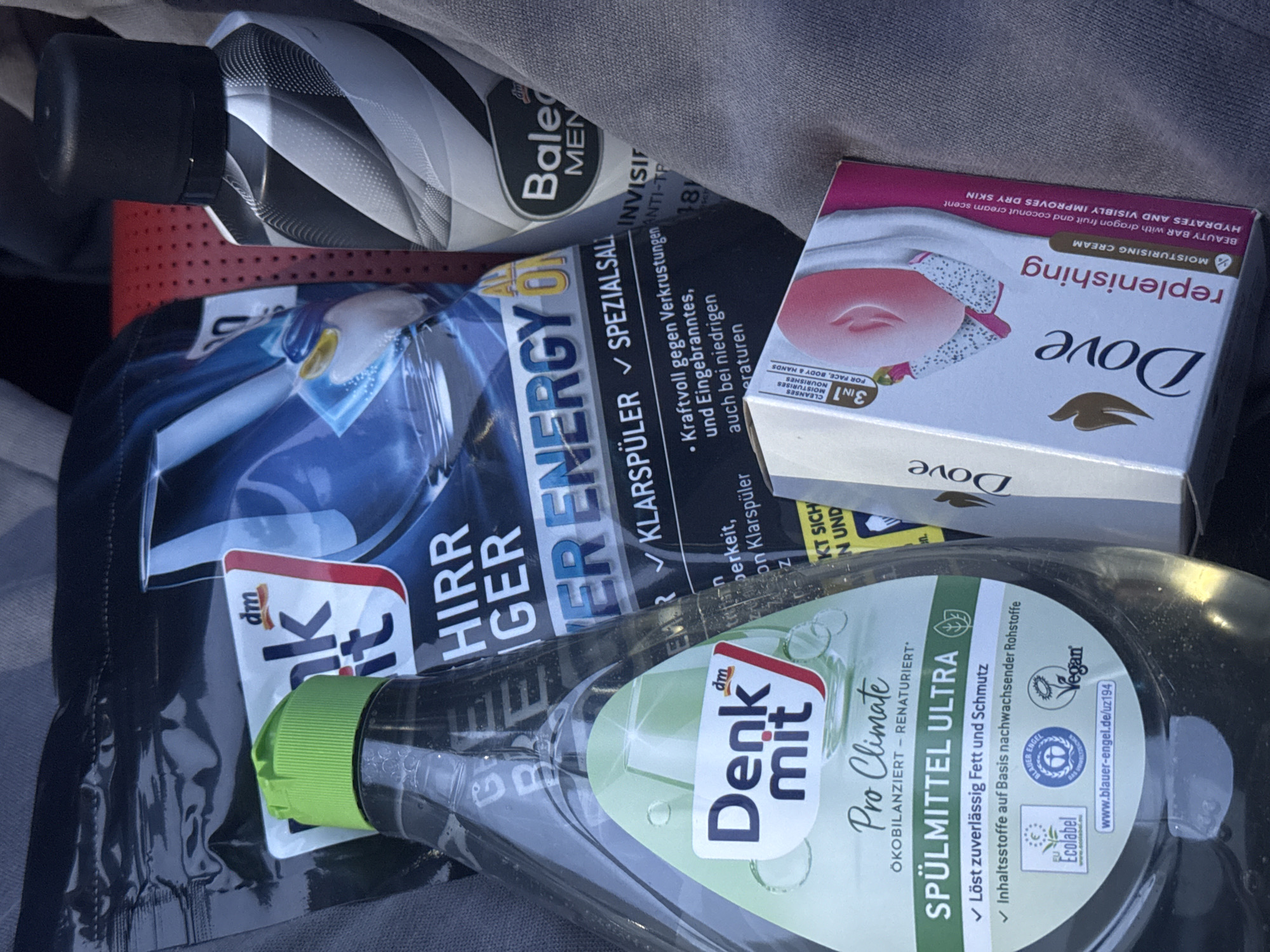 The image is a close-up shot of a collection of household and personal care products. Visible products include: a bottle of "Denk mit Pro Climate SPÜLMITTEL ULTRA" dish soap, a package of "dm Denk mit HIRRIGER ENERGY ON" dishwasher rinse aid, a Dove "replenishing" moisturizing cream beauty bar, and a bottle of "Bale MEN" hair care product. The presence of the "dm" brand indicates these products are from the German drugstore chain, which has branches across Europe. The text on the labels is in German. The background and surroundings are unclear, making it impossible to determine if the photo was taken in Sofia, Bulgaria. There is no visible information to discern if this is in Sofia, Bulgaria.
