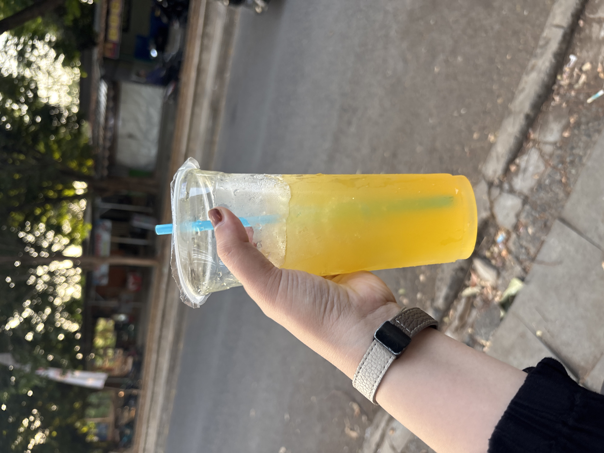 An individual's hand, adorned with a light-colored, textured watchband, firmly grips a transparent plastic cup filled with a bright yellow beverage. A blue straw extends from the cup, partially submerged in the liquid. The cup appears to be sweating, indicating it holds a cold drink. The background is blurred but suggests an outdoor urban environment, likely a street or sidewalk in Cirebon, Indonesia. Trees with dappled sunlight filtering through their leaves line the background, along with indistinct structures that could be storefronts or stalls. A paved road is visible, with hints of parked vehicles further in the distance. The overall scene conveys a sense of casual enjoyment of a refreshing drink in a public area.