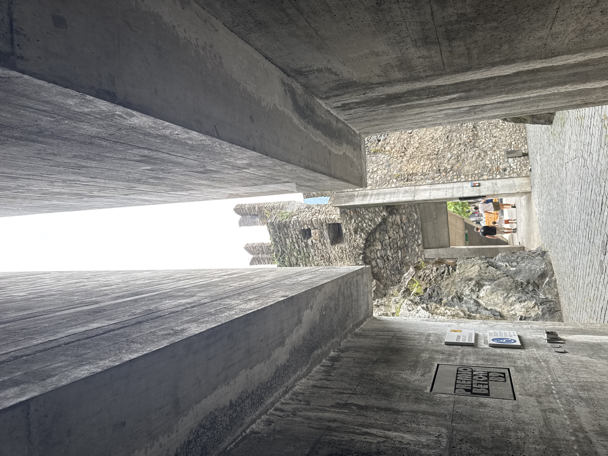 The image shows a pathway leading towards a stone castle tower, framed by two imposing, modern concrete structures. The perspective is from below, looking upwards through a narrow opening between the concrete walls. On the left concrete wall, a rectangular plaque with the text "PREMIO BETON 89" is visible, along with smaller signage. A cobblestone pathway leads into the distance, where two adults and a child are walking away from the camera. The castle tower, constructed of rough stone, has battlements and visible stonework. The environment appears to be an outdoor public space, possibly a historical site or park, in Bellinzona, Switzerland, given the architectural style and the mention of "PREMIO BETON," which relates to concrete awards. The lighting suggests an overcast or diffused daylight.