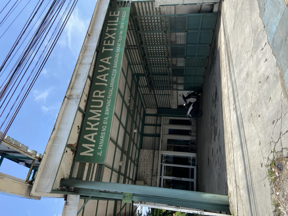 The image shows the exterior of a business in Padalarang, Indonesia, located at coordinates -6.846 latitude and 107.498 longitude. The business is a textile company named "Makmur Jaya Textile", which is located on JL. Panaris No. 61A, Simpang Padalarang K.B, Bandung Barat. The business has a sign above the entrance with its name and contact information. There is a motorbike parked in front of the building, and several power lines are visible in the sky. The building is made of concrete and has a green roof. The entrance to the building is through a set of double doors. The image was taken during the day. The sky is clear and blue. There are no people in the image.