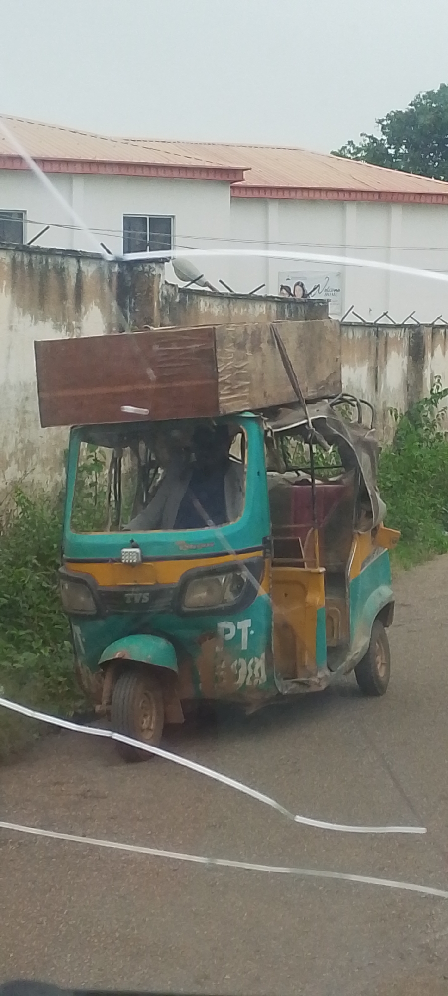This media file shows a light blue and yellow auto-rickshaw, commonly known as a "keke," parked on an asphalt road. The keke is heavily laden with cargo. Two large wooden planks are stacked on top of the keke's canopy, extending significantly beyond its width. The keke itself appears aged and somewhat worn, with visible rust and dirt. White plastic piping runs across the image, seemingly in the foreground and background, possibly indicating construction or maintenance work.
In the background, a light-colored building with a reddish-brown roof is visible. A concrete wall with a textured surface and a jagged metal fence runs in front of the building. Overgrown green vegetation can be seen on the right side of the frame, bordering the road. The sky is overcast, suggesting a cloudy or possibly rainy day. The lighting is diffused, and there are no strong shadows, consistent with a cloudy sky.
On the side of the keke, the text "PT" is visible, followed by what appears to be a number "98" or similar characters, rendered in a white font. The overall impression is a scene of everyday transportation and cargo in a semi-urban or rural environment, possibly in Gura Top, Nigeria, given the location context. The activity depicted is the parking of the loaded keke, implying it is either stopped for a break or about to depart. There are no people clearly visible within the keke or the immediate surroundings, though a figure can be dimly seen inside the driver's cabin.