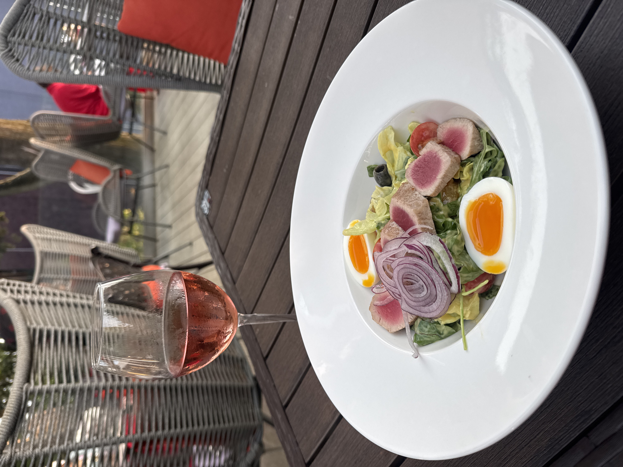 The image depicts a close-up view of a salad dish and a glass of rosé wine on a wooden table. The salad, served in a white bowl, contains seared tuna, hard-boiled eggs, red onion rings, and greens. In the background, several wicker chairs with red cushions are visible, suggesting an outdoor seating area, possibly at a restaurant or cafe. The scene is set in Sochi, Russia.