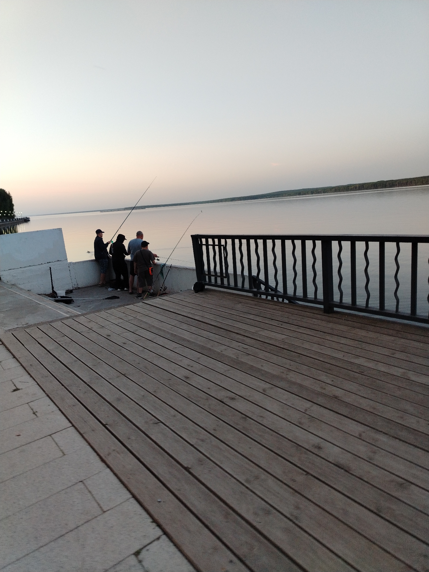 Four people are fishing from a concrete and wooden pier. Fishing rods are visible extending over the water. The pier has a decorative metal railing. The water is calm, and trees line the opposite shore. The scene is at dusk or dawn, with a light-colored sky. The location is Chaikovsky, Russia.