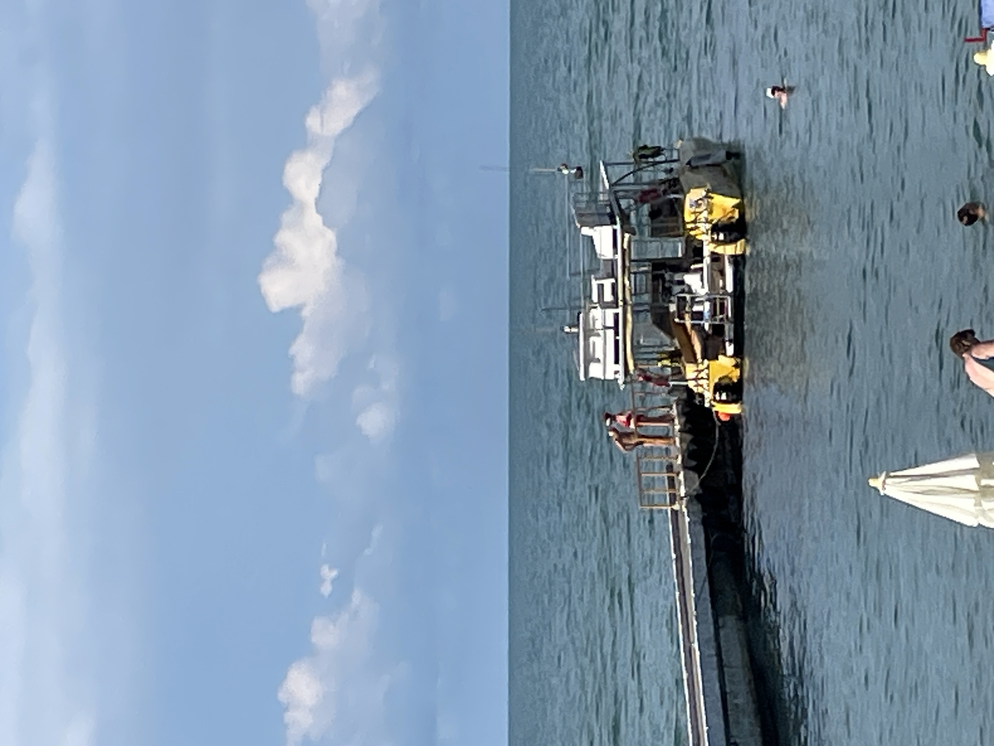 The image captures a lively scene on the water, likely in Sochi, Russia. The primary subjects are several people enjoying a day by the sea. Some are gathered on a floating structure, possibly a boat or a pontoon, which is yellow and white. Others are in the water, swimming or wading. One person is visible in the distance, appearing to be swimming.
The setting is a clear, bright day with a blue sky and scattered white clouds. The water is a deep blue-green and appears calm, with gentle ripples. The sun is high, suggesting it's likely midday. A white umbrella is partially visible on the right side of the frame, hinting at a beach or recreational area nearby.
There are no visible signs or text in the image. The overall mood appears to be one of leisure and enjoyment, characteristic of a summer day spent by the sea. The image focuses on the recreational aspect of the location, with people actively engaged in water activities. The floating structure with people on it adds a dynamic element to the scene. The presence of multiple individuals in the water and on the structure indicates a social gathering or a popular spot for water activities.