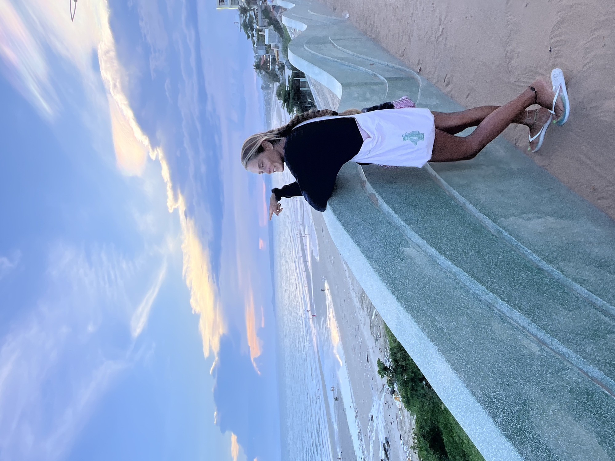 The image shows a woman standing on a sandy path next to a textured green concrete wall. She is wearing a black long-sleeved shirt, a white tote bag with a cartoon bear graphic, a pink and white plaid skirt, and flip-flops. Her left arm is resting on the wall, and she is pointing towards the sea with her right index finger. She is looking to her right, away from the camera.
In the background, there is a beach with several people scattered along the shoreline and in the water. The sea is calm, and the sky above is filled with dramatic clouds tinged with orange and pink hues, indicating either sunrise or sunset. Buildings and palm trees are visible along the coastline in the distance. The scene is set in Mũi Né, Vietnam, a coastal town known for its beaches and sand dunes.