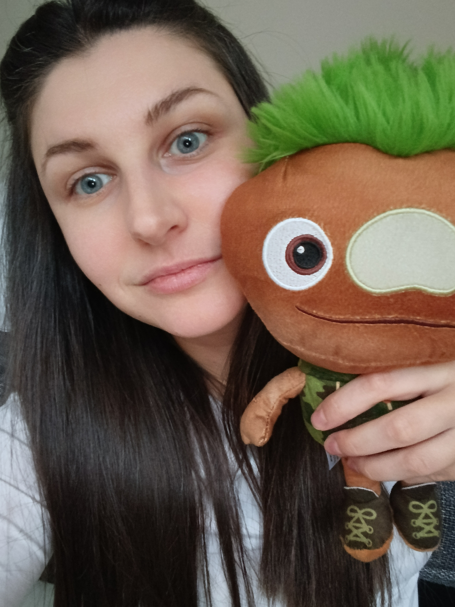 A woman with long, dark hair and blue eyes is holding a plush toy. The toy is brown with green hair, wearing a camouflage vest and brown shoes. The background is a neutral color. The location is Unknown, Unknown.