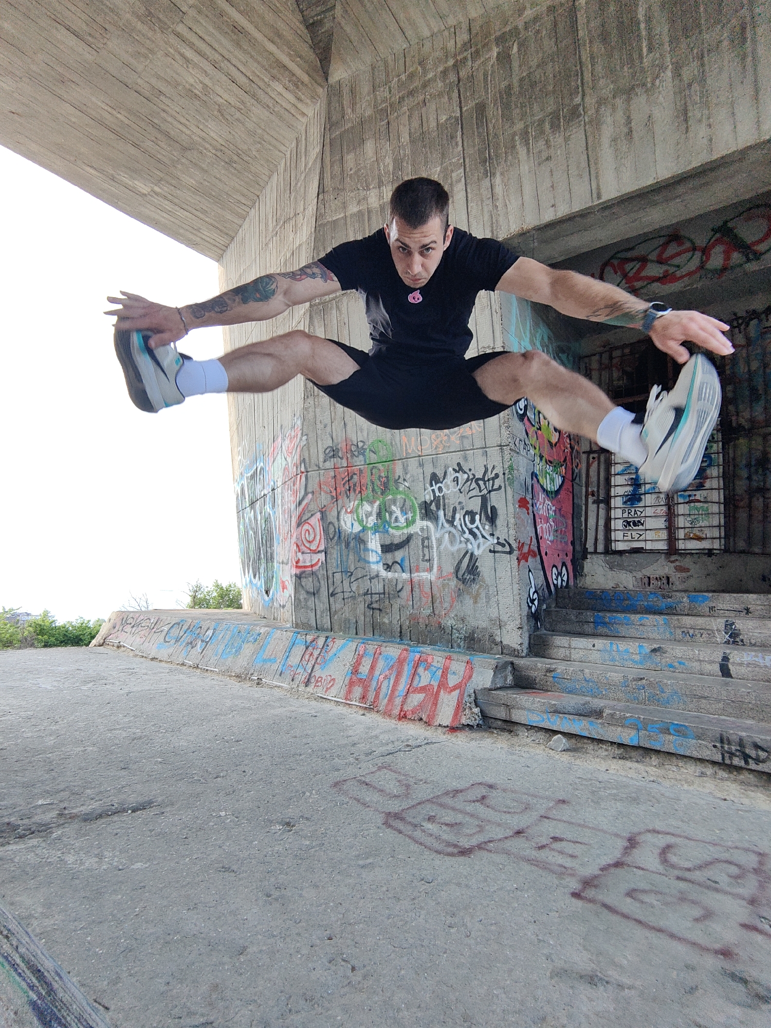 A man is performing a split jump in front of a concrete wall covered in graffiti. The man is wearing a black t-shirt, black shorts, white socks, and light-colored running shoes. The background shows a concrete structure, possibly a bridge or underpass, with graffiti on the walls and steps. The location is likely within an urban area in Varna, Bulgaria, given the setting and graffiti style. The lighting suggests it is daytime.